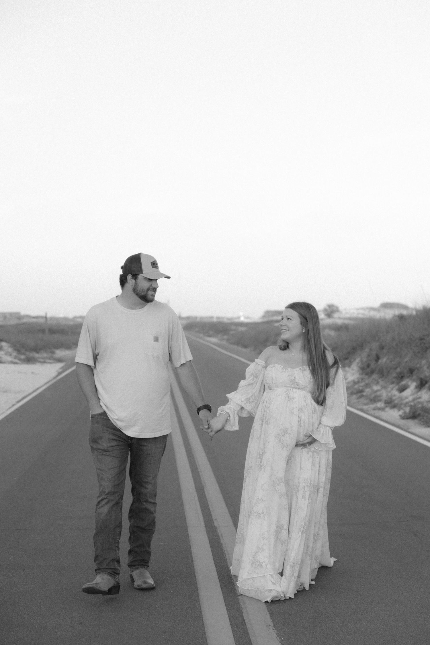Black and white photo of a pregnant couple holding hands on a road, woman in lace gown, man in cap and jeans.