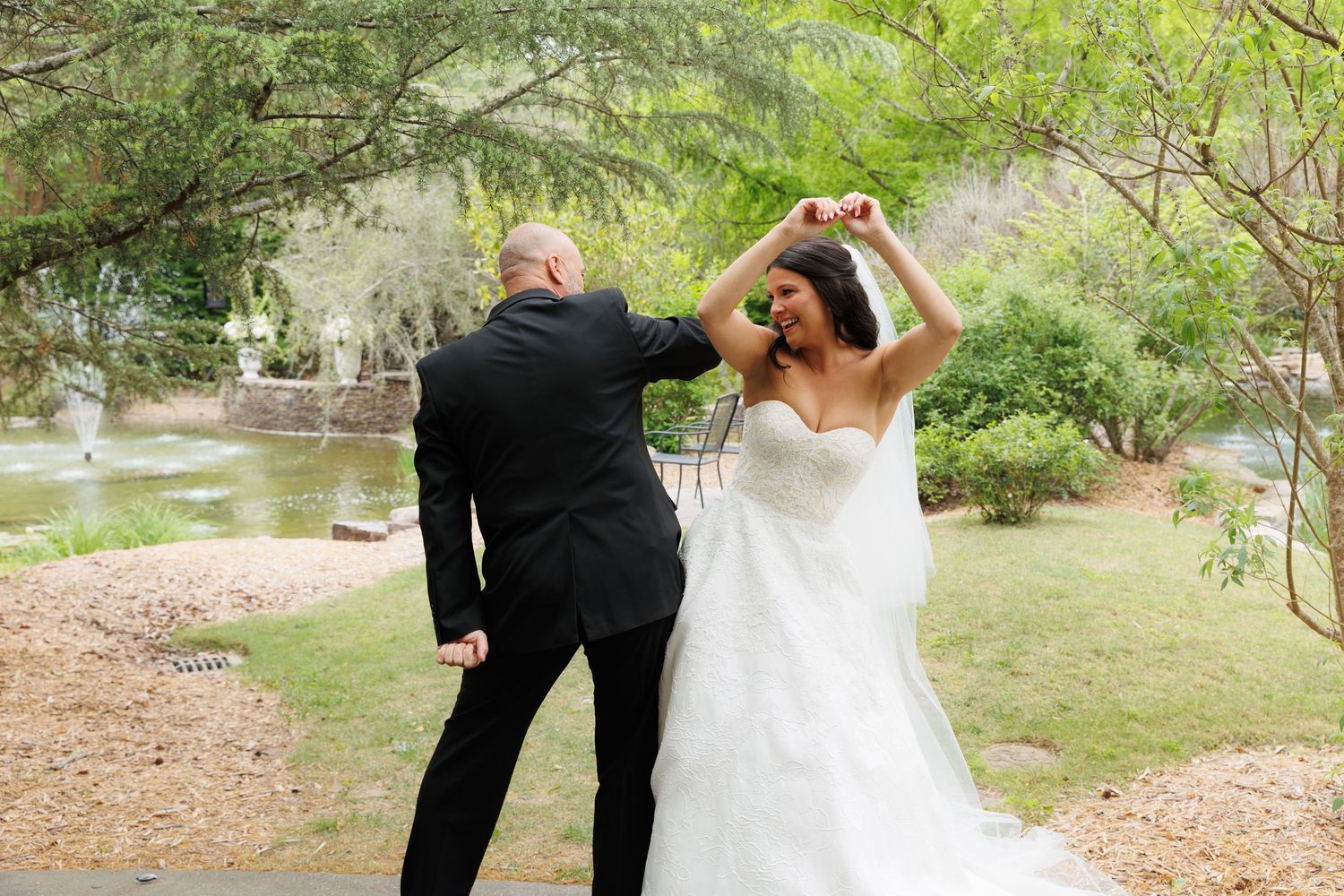 Bride in white strapless gown being twirled by groom in black suit outdoors in lush garden setting.