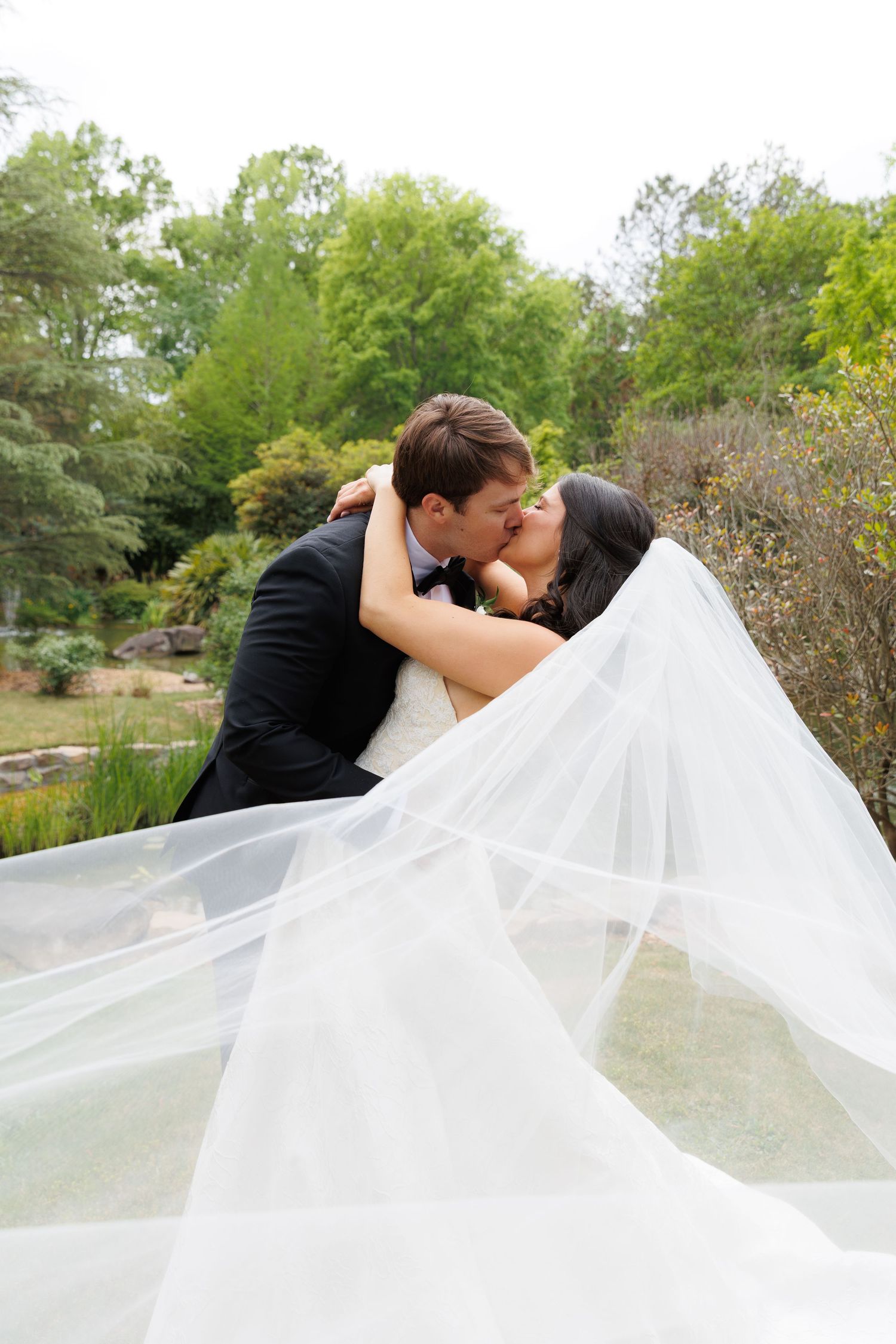 Bride and groom share a romantic kiss outdoors with wedding veil flowing in the breeze amid lush green garden backdrop.