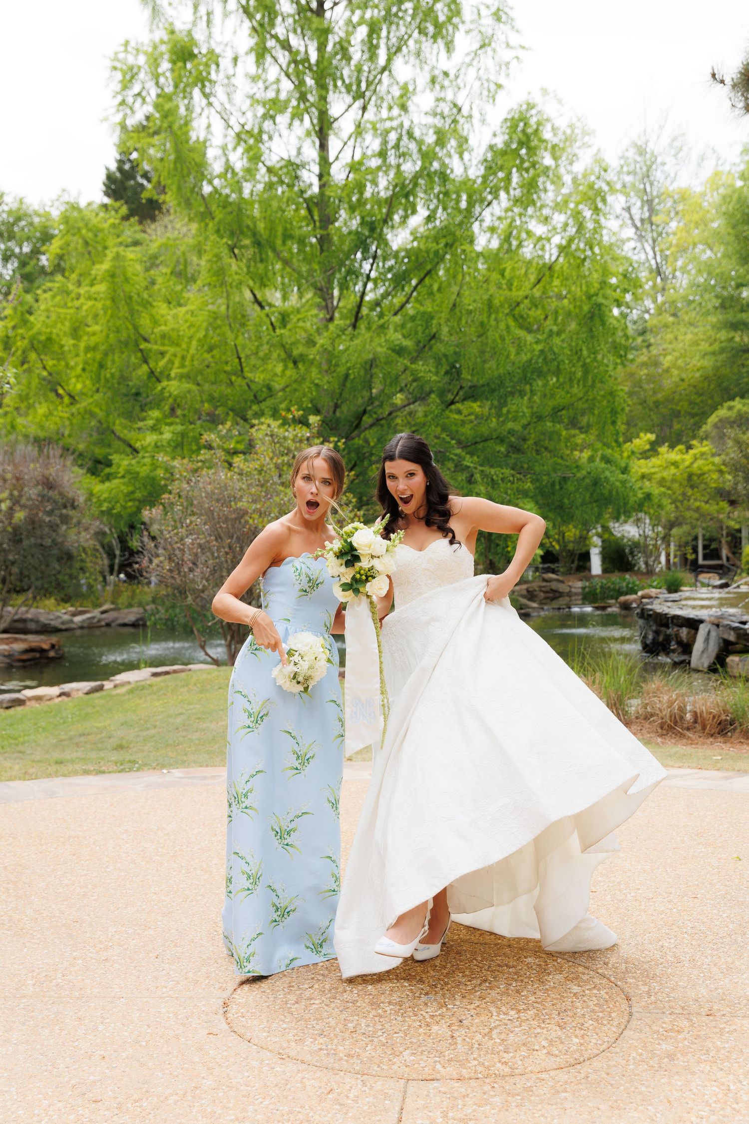 Bride in white gown and bridesmaid in blue floral dress pose playfully with bouquets in an outdoor garden setting.