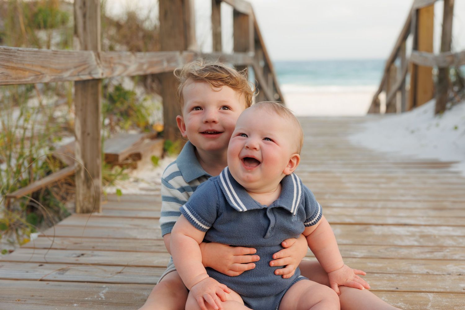 Two happy young brothers sitting on a beach boardwalk, smiling with ocean in background.