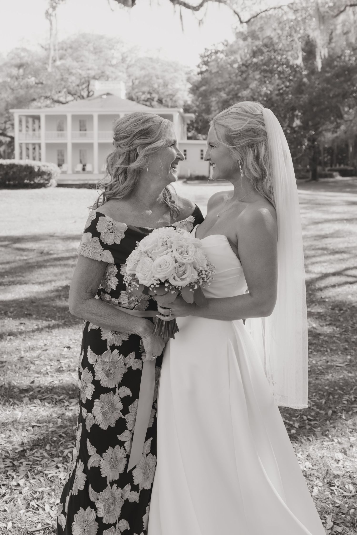 Black and white photo of a bride in white gown and veil holding bouquet, smiling with woman in floral dress outdoors.