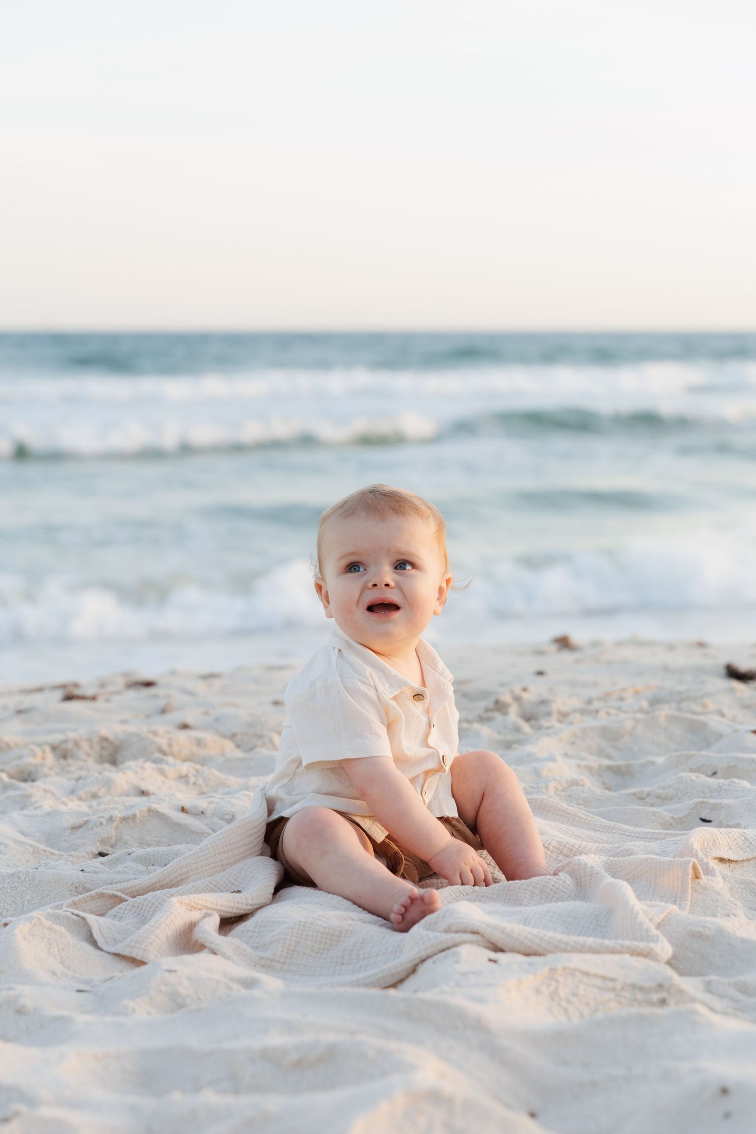 A happy baby sits on a white sandy beach blanket with ocean waves in the background.