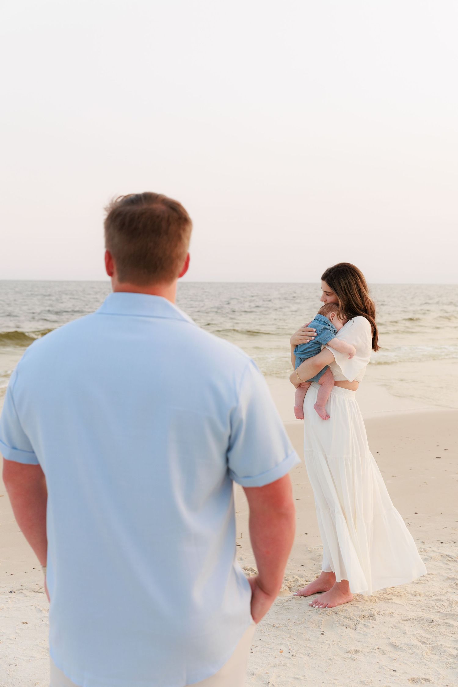 A man watches a woman in a white dress holding a baby on a serene beach with ocean waves in the background.