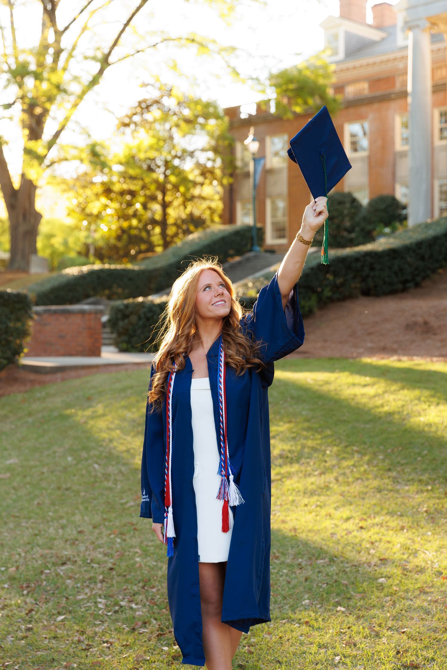 Smiling graduate in navy cap and gown raises mortarboard on sunny campus lawn.