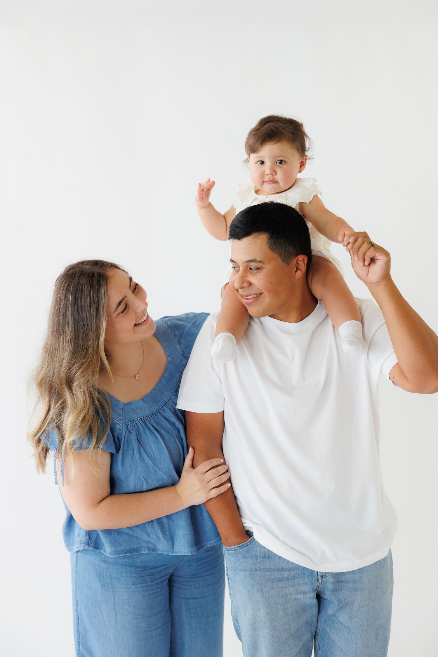 Happy family portrait with mom, dad, and baby sitting on father's shoulders against white background.