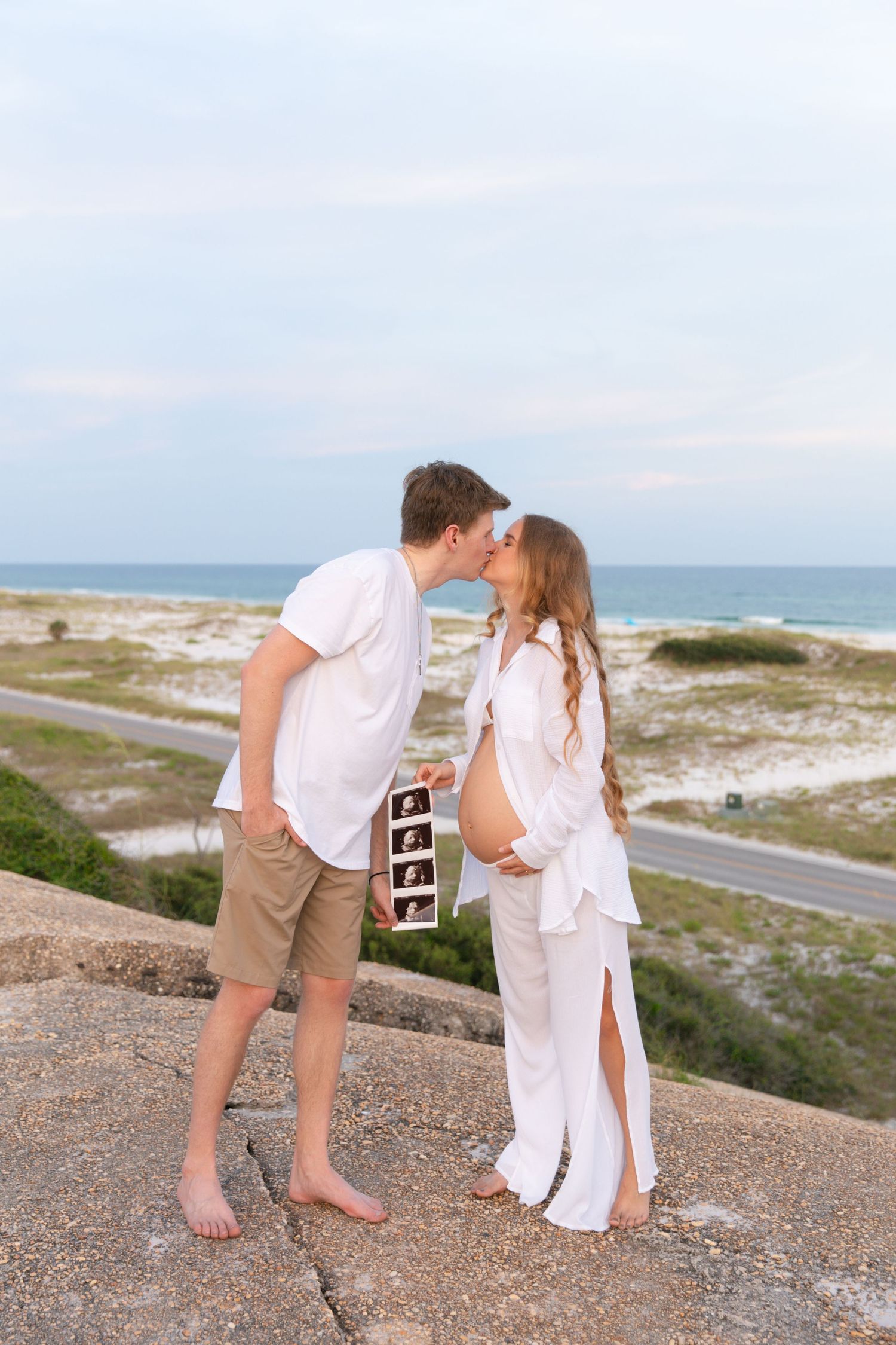 A couple shares a romantic moment on a coastal overlook while holding a series of ultrasound photos.