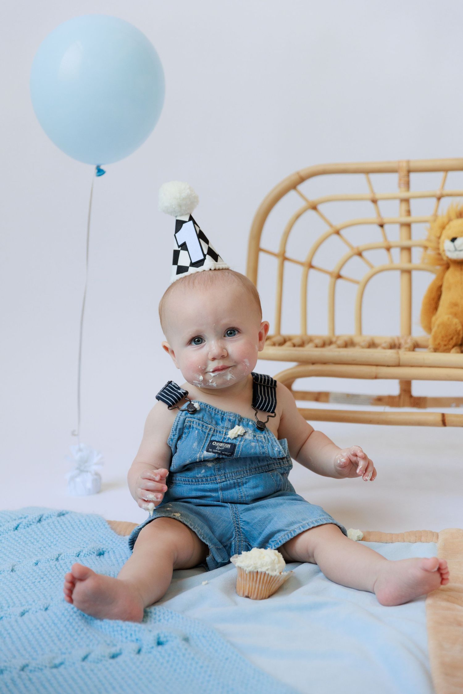 Cozy nursery setting with blue balloon, rattan furniture, and knit blanket in soft pastel tones.