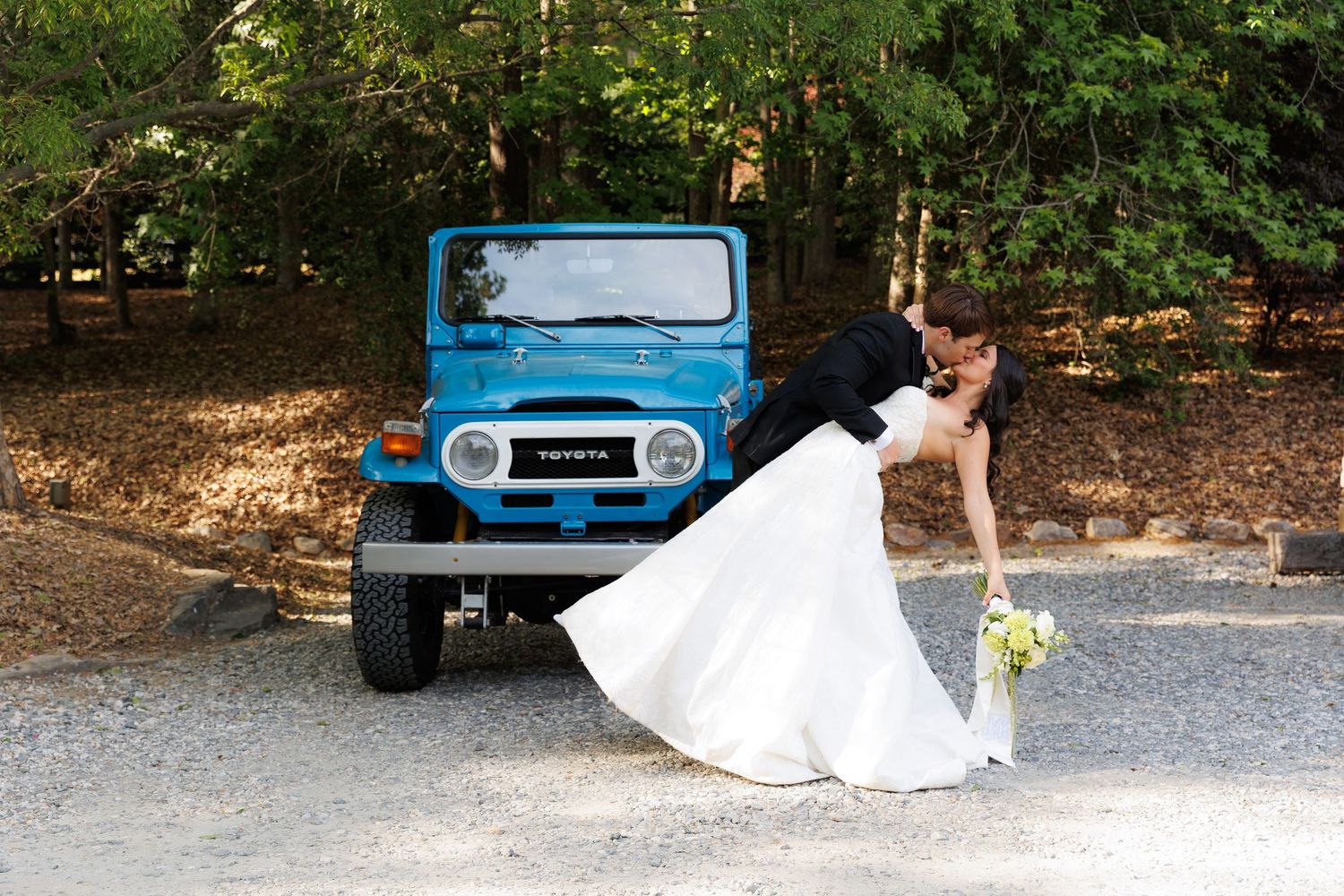 Bride and groom sharing a romantic dip kiss in front of a vintage blue Toyota FJ40 outdoors.