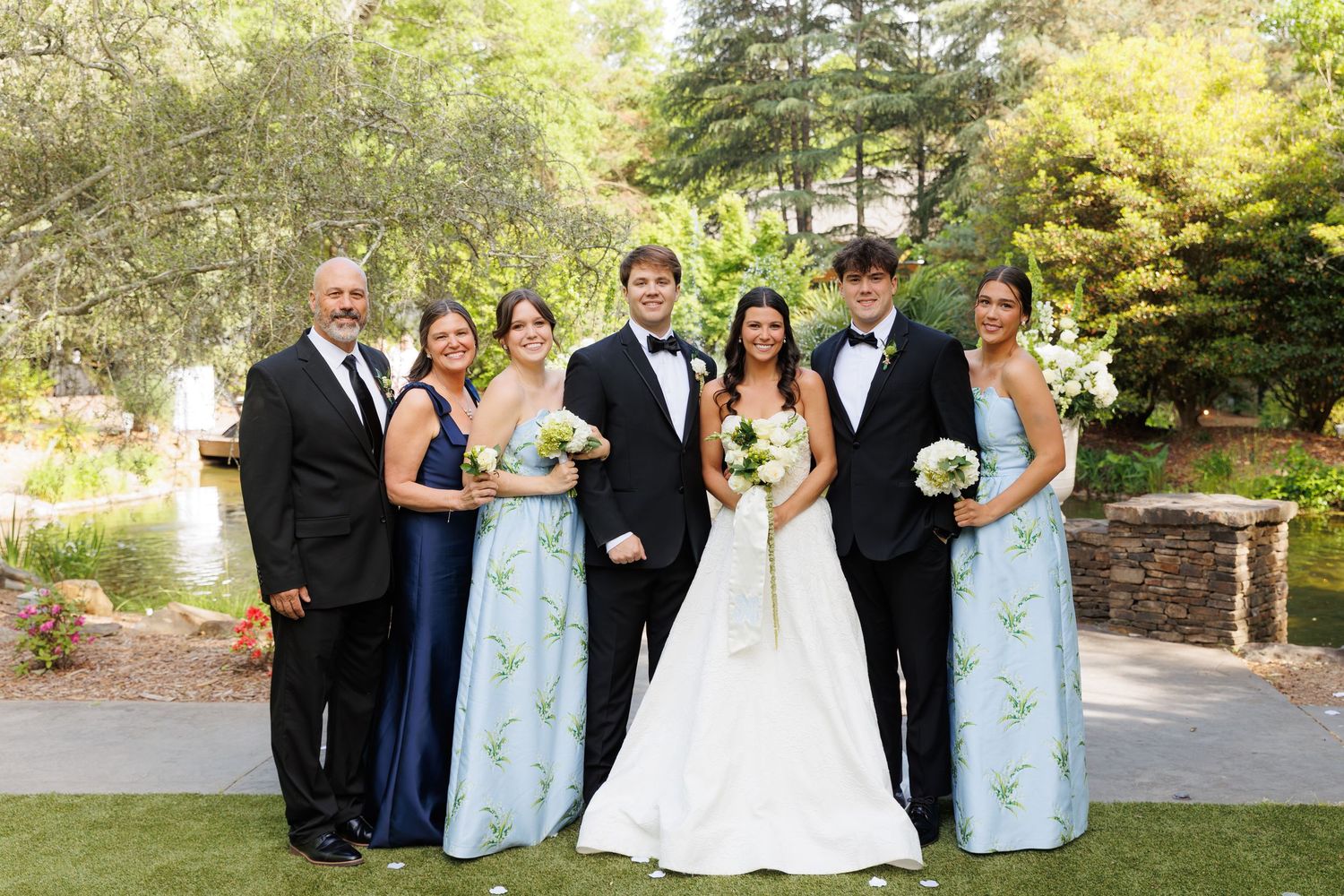 Wedding party poses outdoors in lush garden, bride in white gown, groom in tuxedo, bridesmaids in blue dresses.