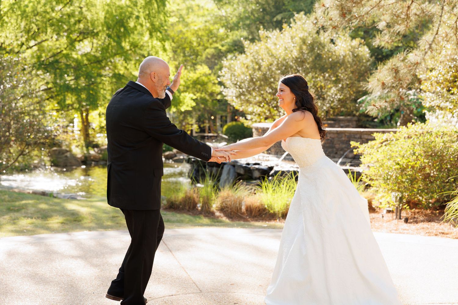 Bride and groom hold hands and spin together outdoors in a sunny garden during their wedding day first look.