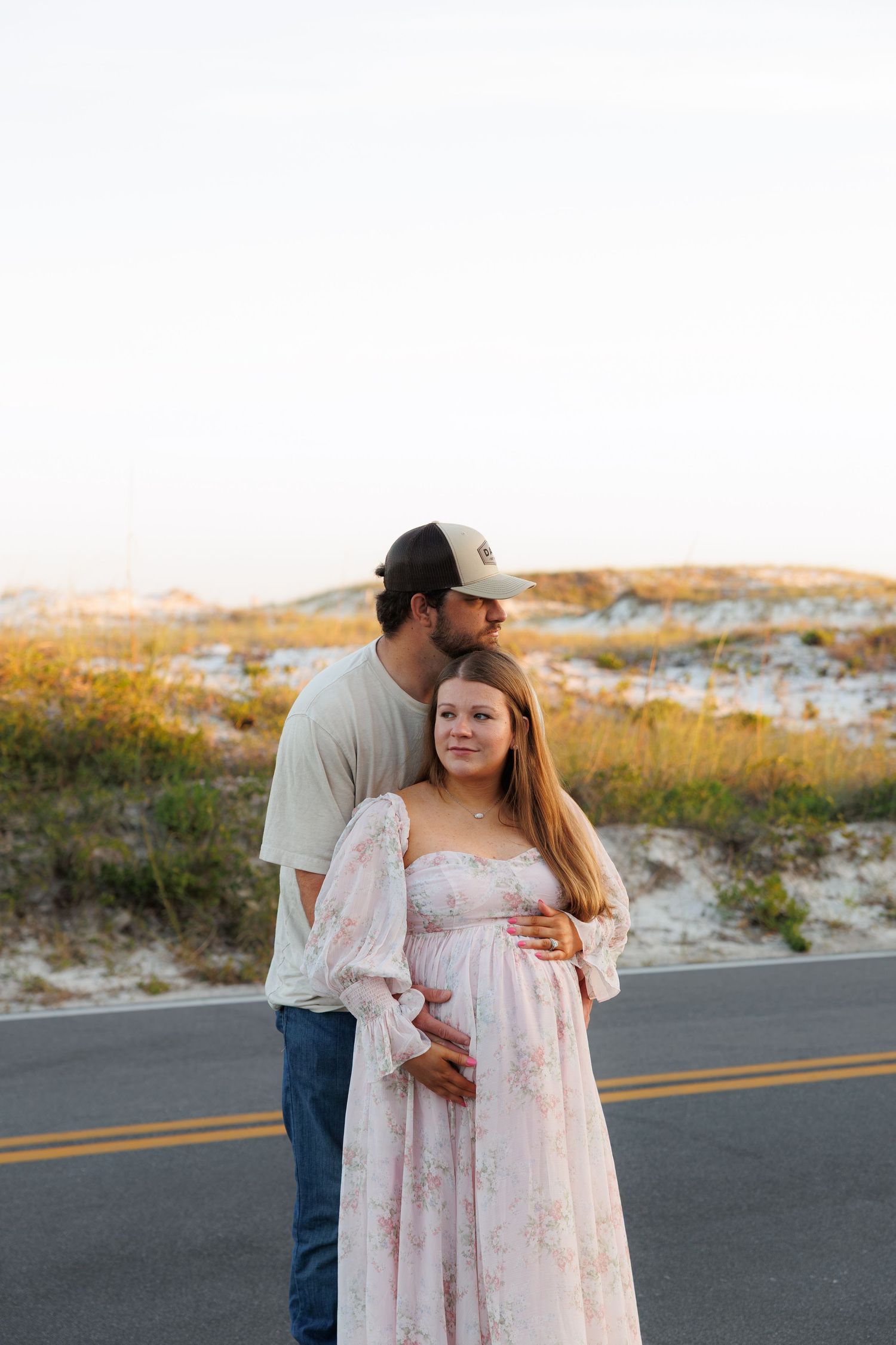 Couple embracing outdoors at sunset, man kissing pregnant woman in floral dress near sand dunes.