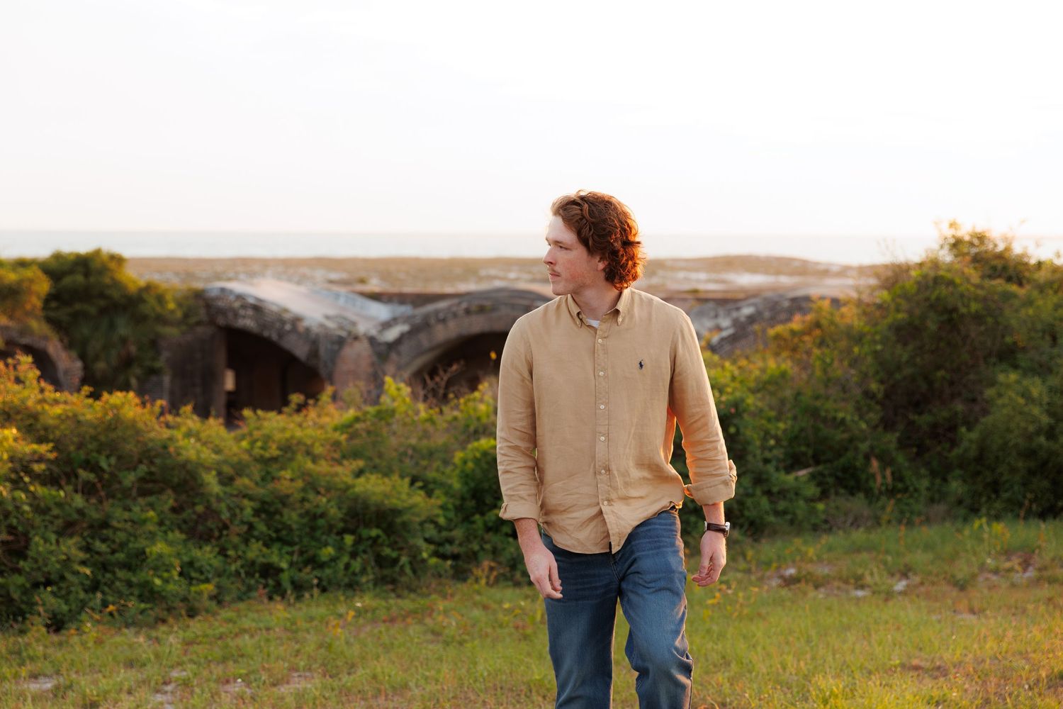 A young man with curly red hair in a tan shirt and jeans stands outdoors near historic stone ruins at golden hour.