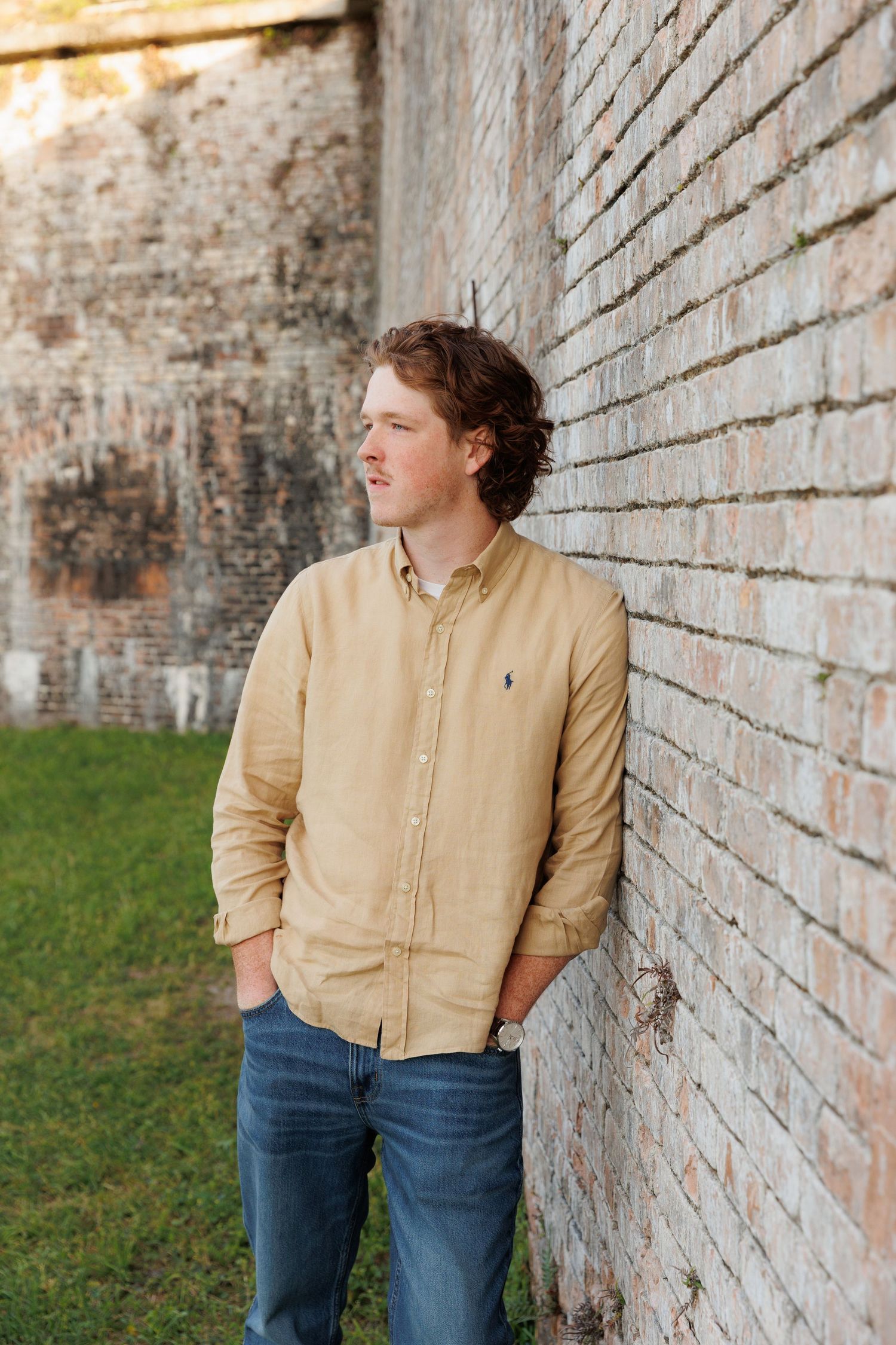 Young man in tan Polo shirt and jeans leaning against rustic brick wall outdoors.