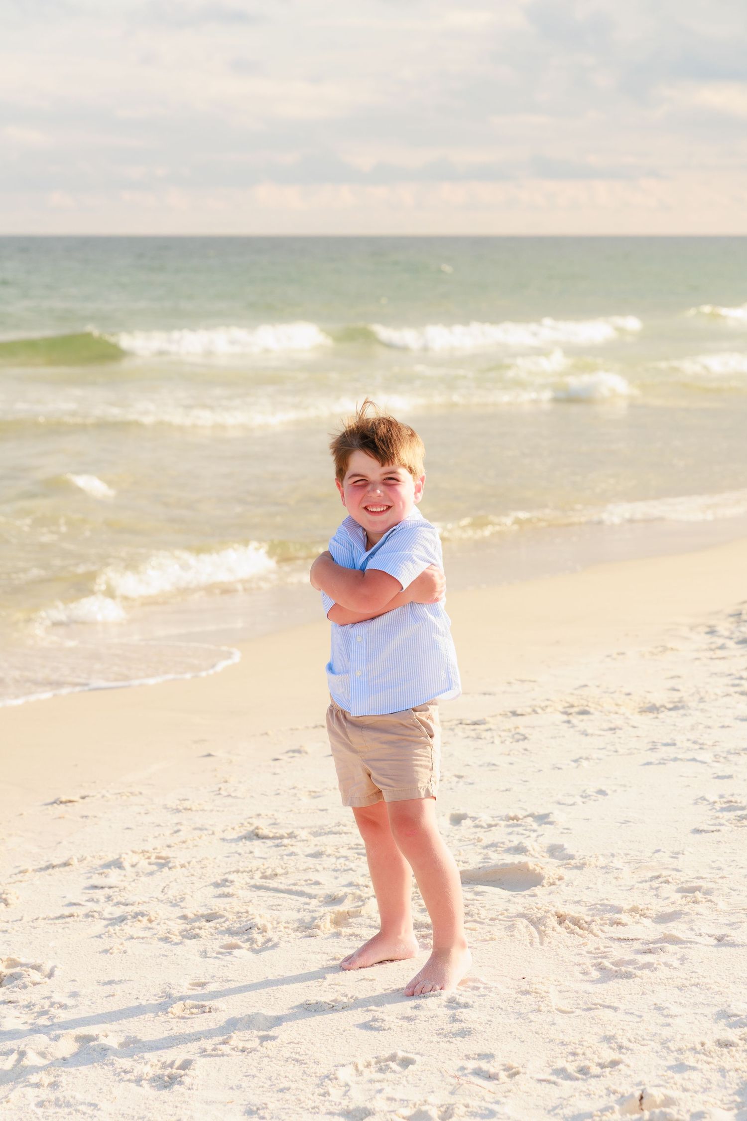 A smiling boy with arms crossed stands barefoot on a sandy beach with ocean waves in the background.