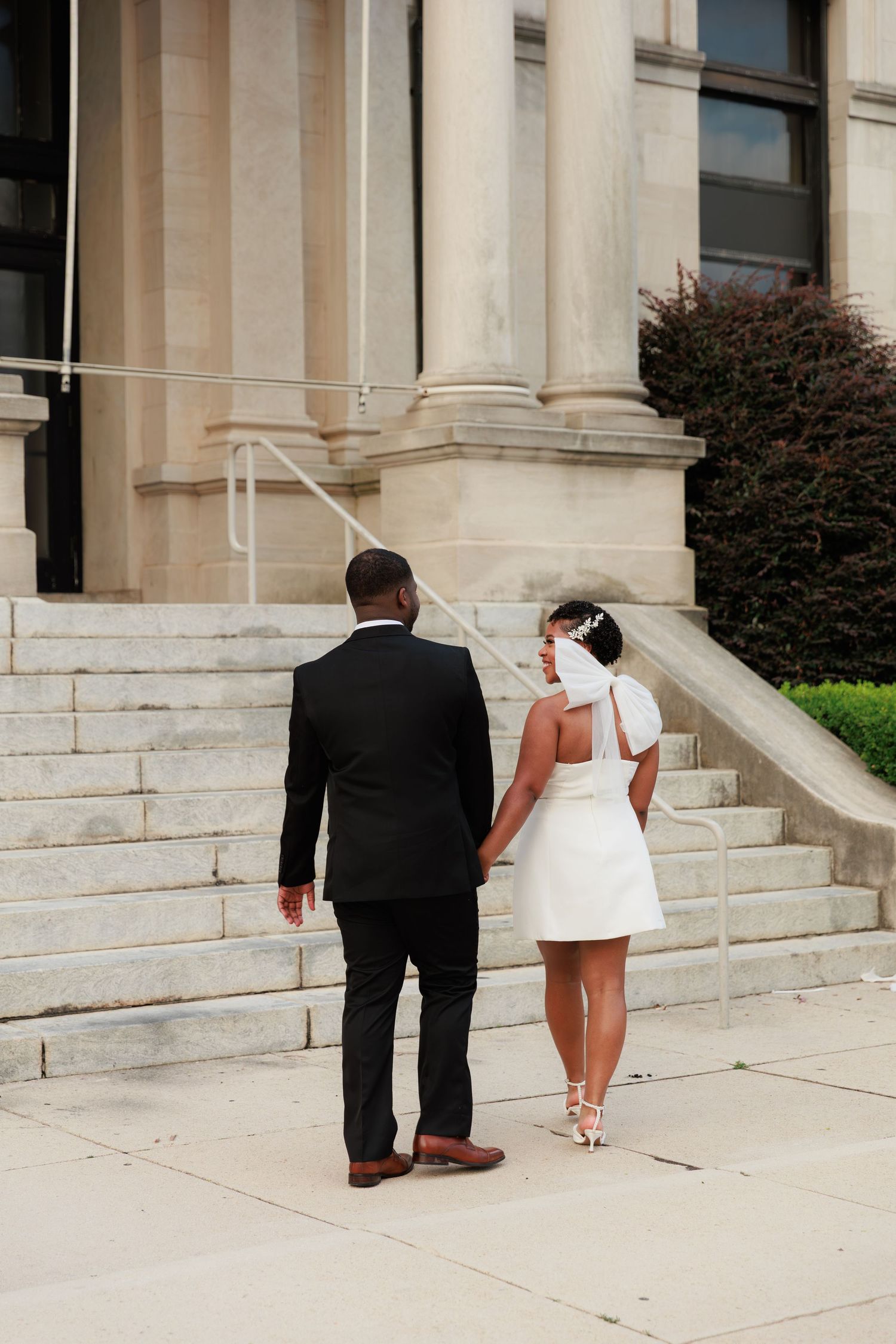 A Black couple in wedding attire holds hands walking toward stone steps of a grand building, bride in short white dress.