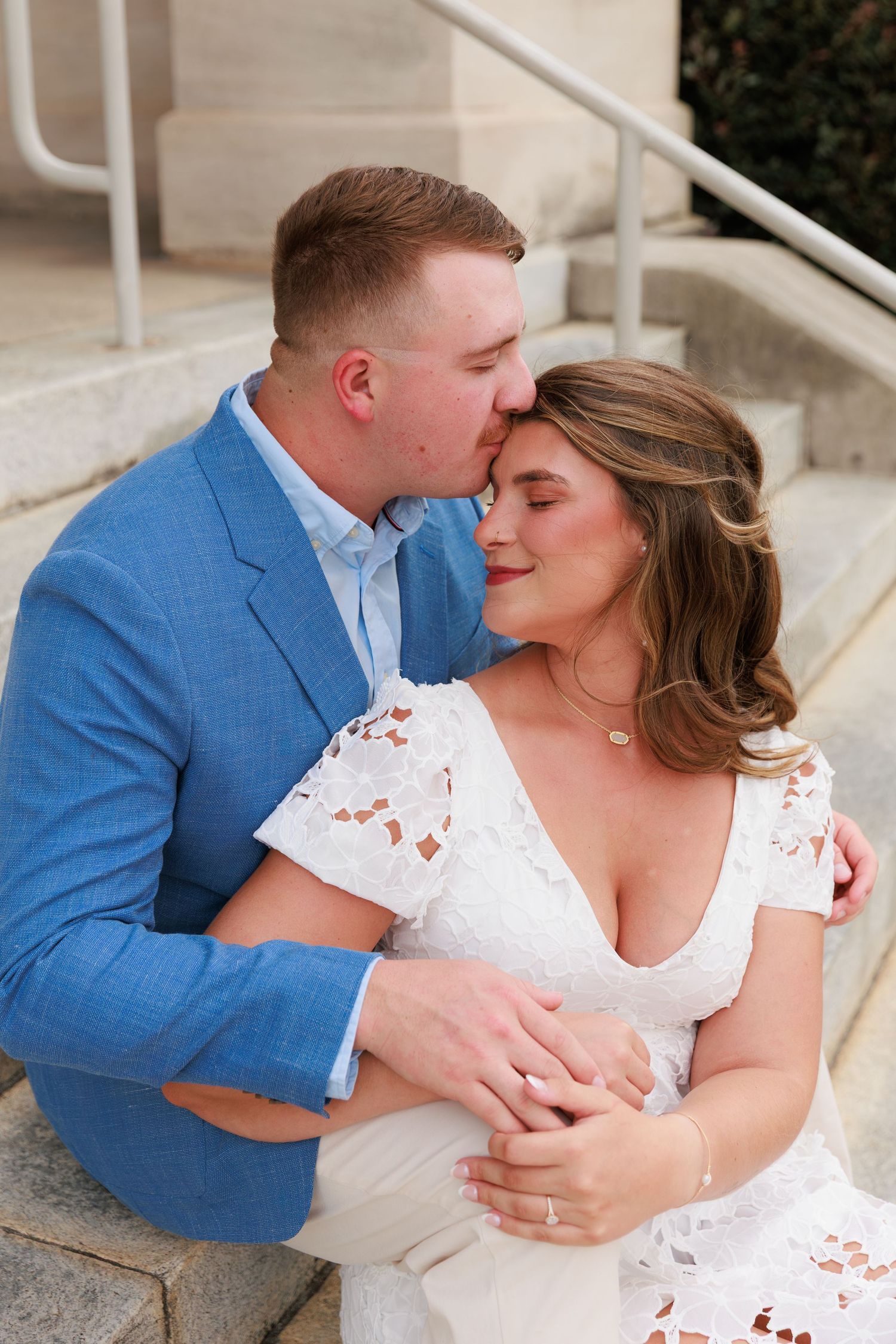 Couple sitting on stone steps, man in blue blazer kissing woman in white lace dress on the forehead.