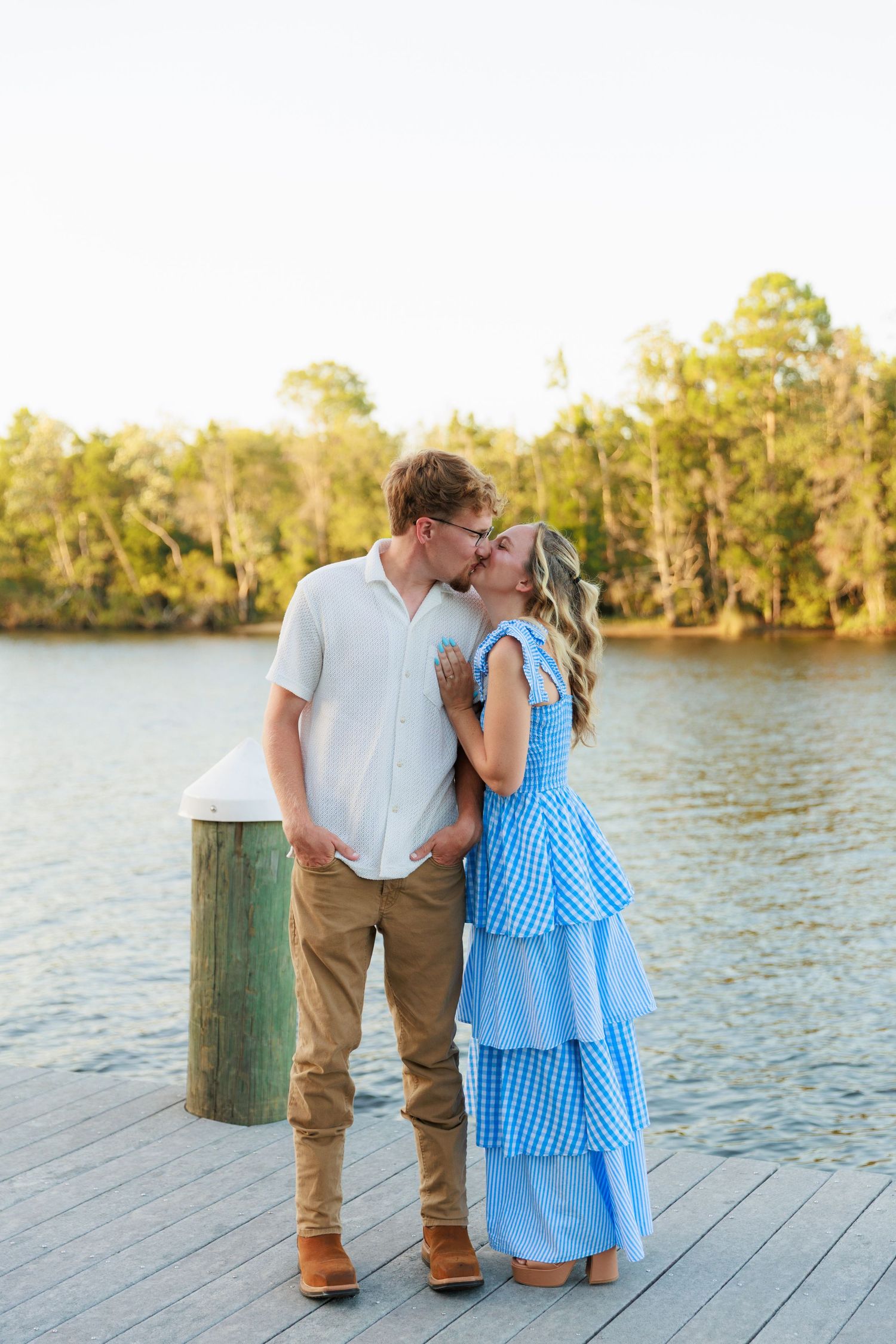 Couple sharing a kiss on a wooden dock by a serene river, woman in blue gingham tiered dress, man in white shirt.