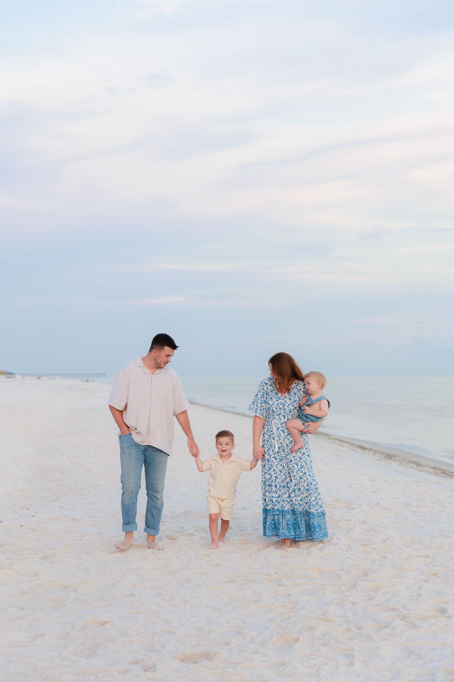 Happy family of four walks on white sand beach at dusk, parents holding hands with toddler and baby.