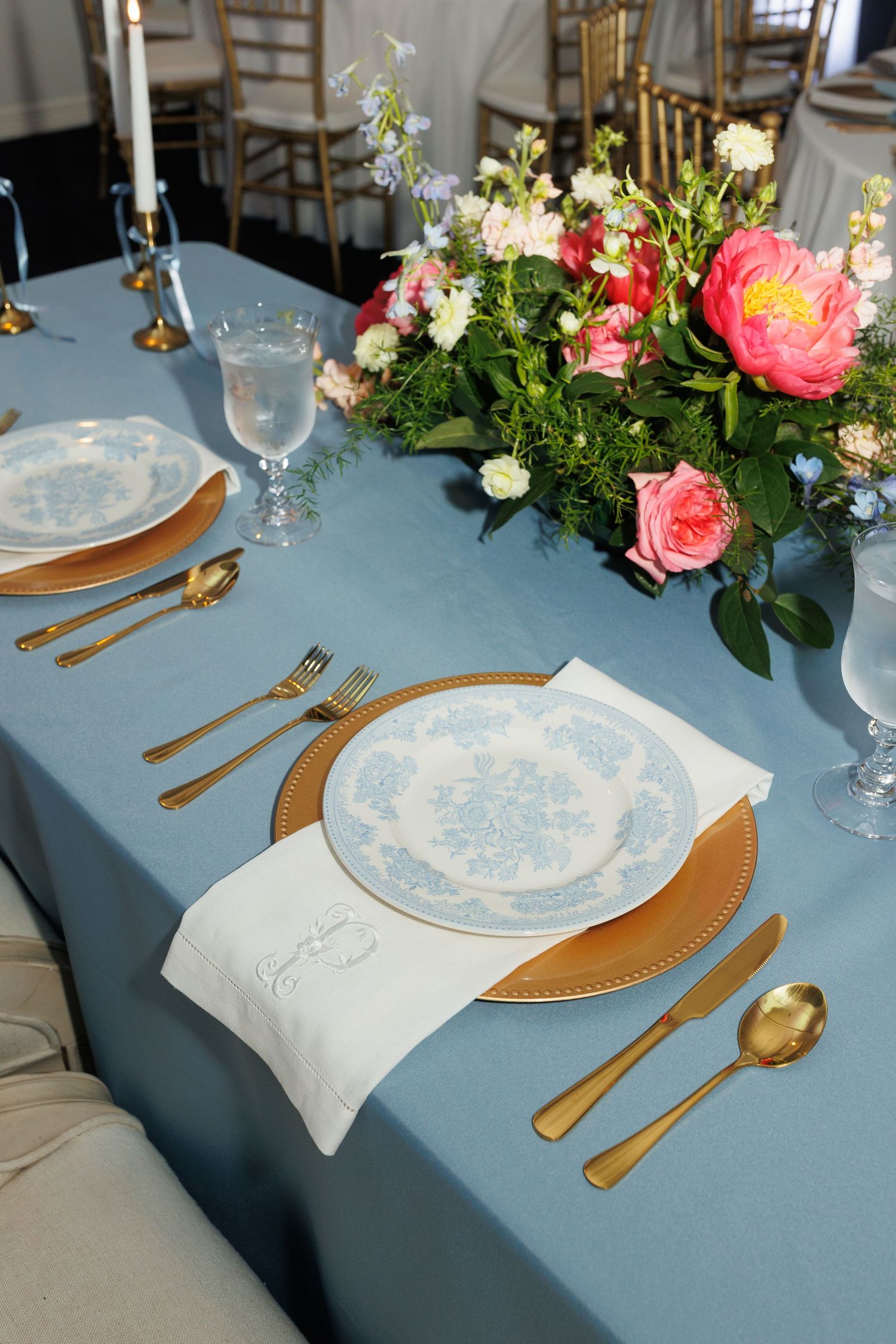 Elegant table settings with blue tablecloth, gold cutlery, white china plates and pink floral centerpieces at a formal event.