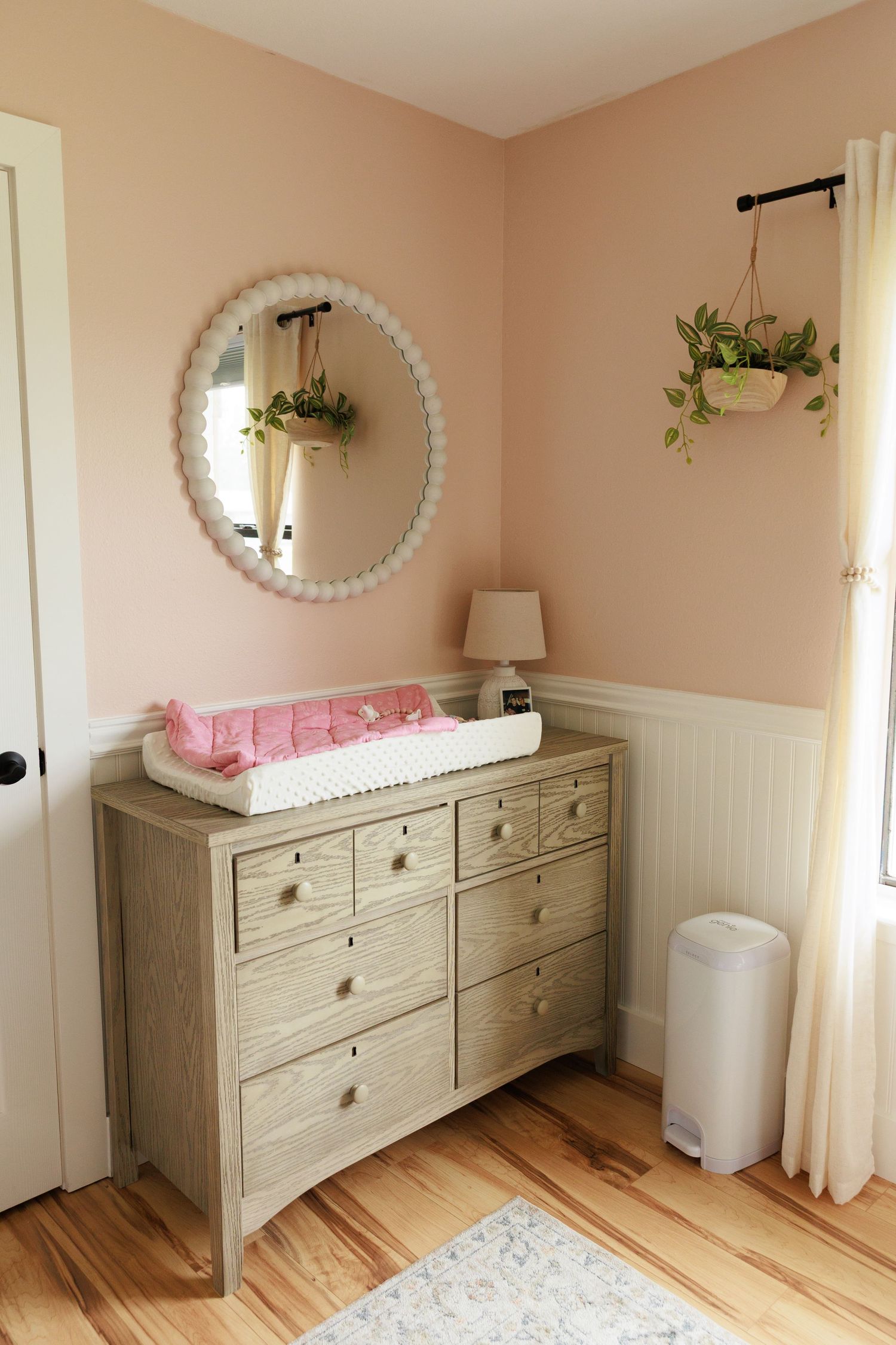 A nursery dresser with changing pad and round mirror, featuring blush walls and hanging plants in a cozy corner.