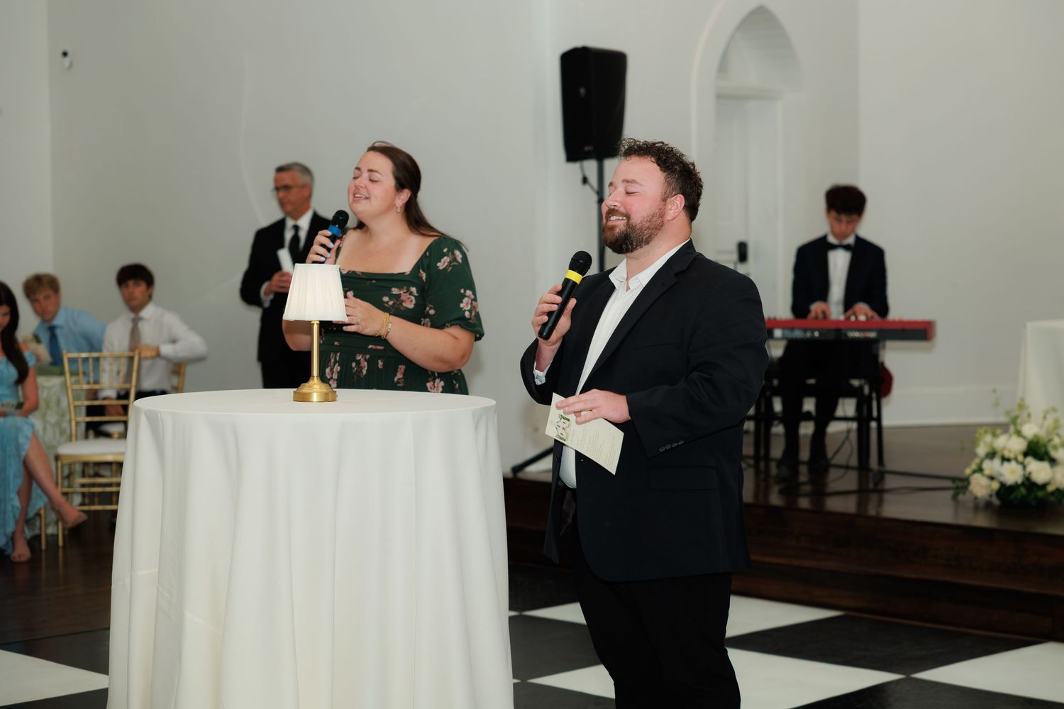 Two wedding MCs speaking into microphones at a white podium in an elegant venue with guests seated behind.