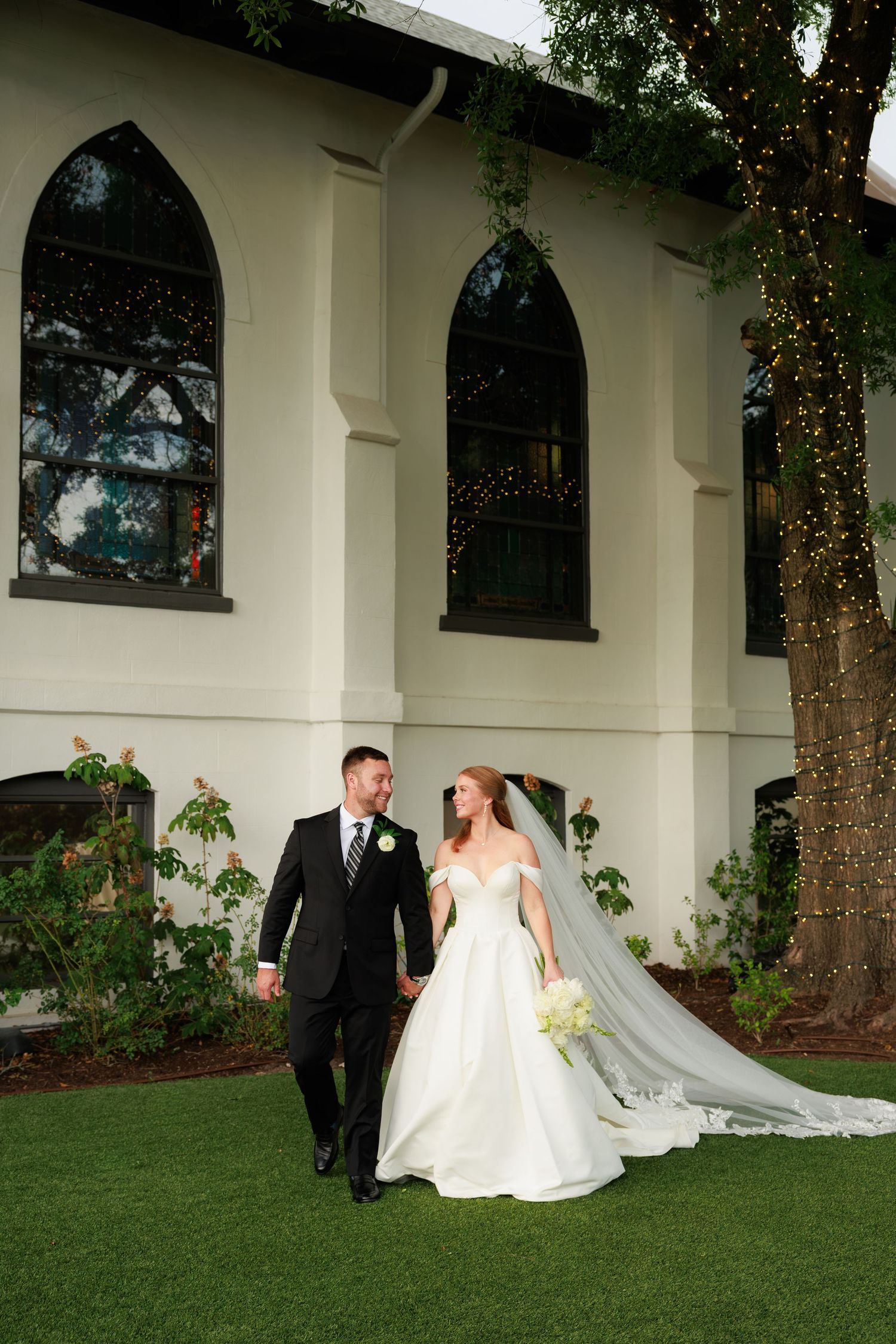 Bride and groom walk hand-in-hand outside white chapel with arched windows and twinkling fairy lights in trees.