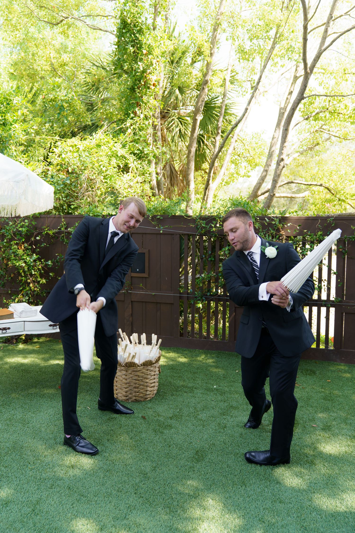 Two groomsmen in black suits playfully swing white fabric items outdoors at a wedding reception on a green lawn.