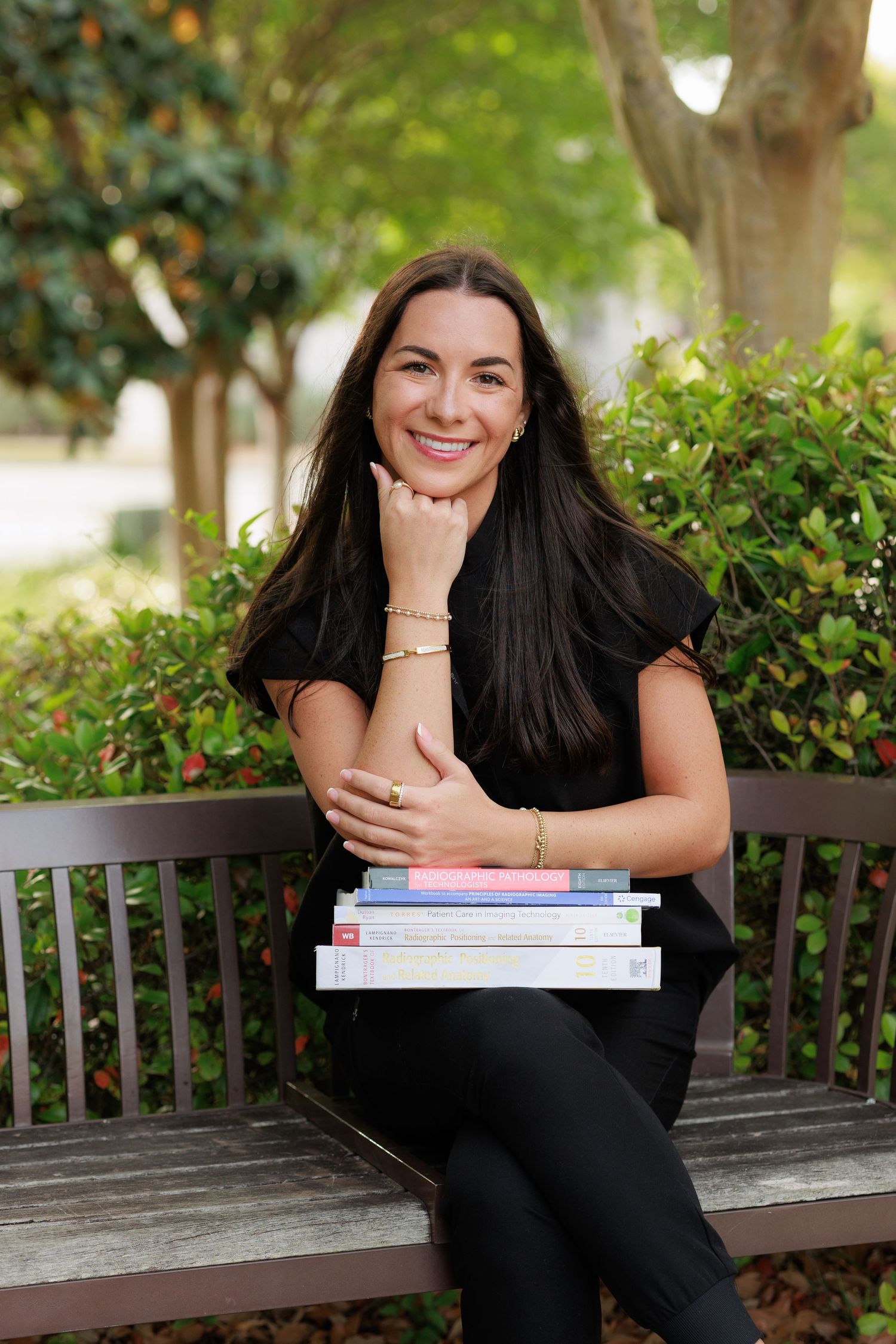 Smiling woman in black outfit sitting on bench with stack of books, surrounded by lush green shrubs outdoors.
