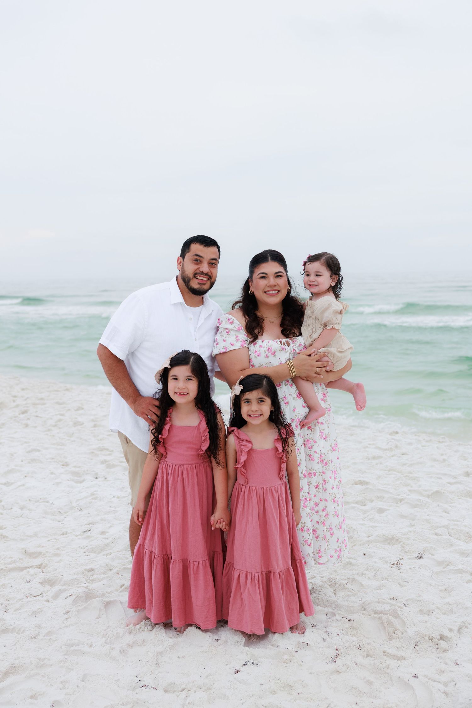 A family of five poses on a white sand beach, girls in pink dresses with parents and baby girl.