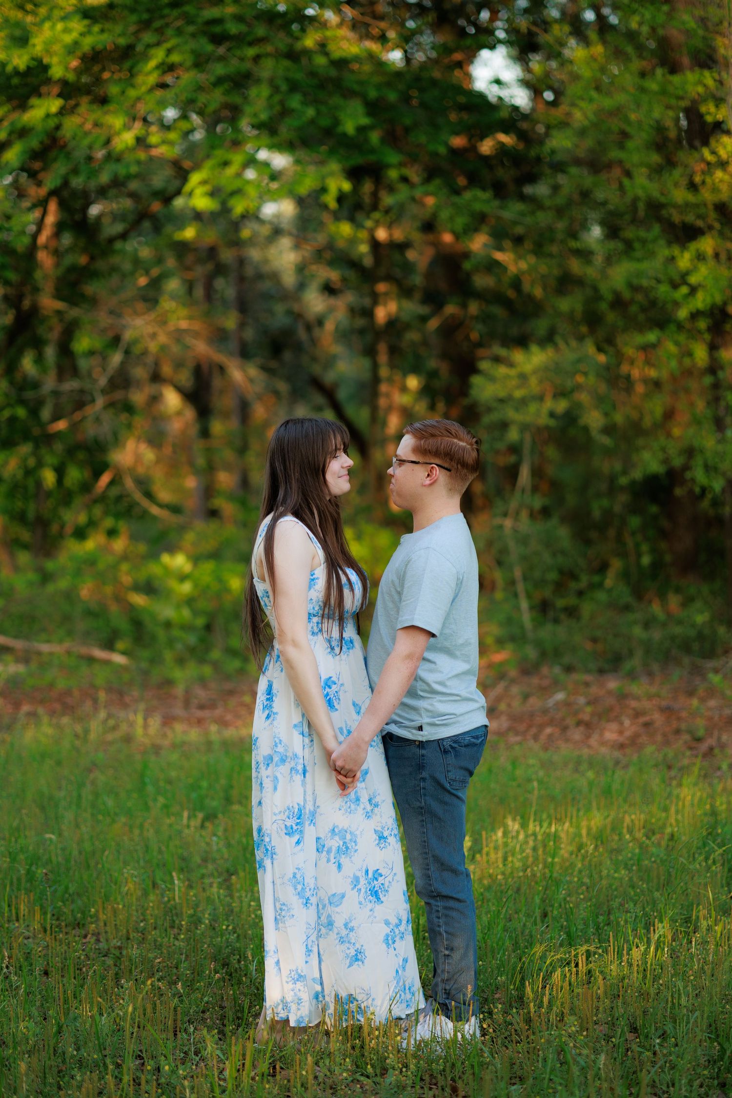 A young couple holds hands and gazes at each other in a lush green meadow, she wearing a blue floral dress.
