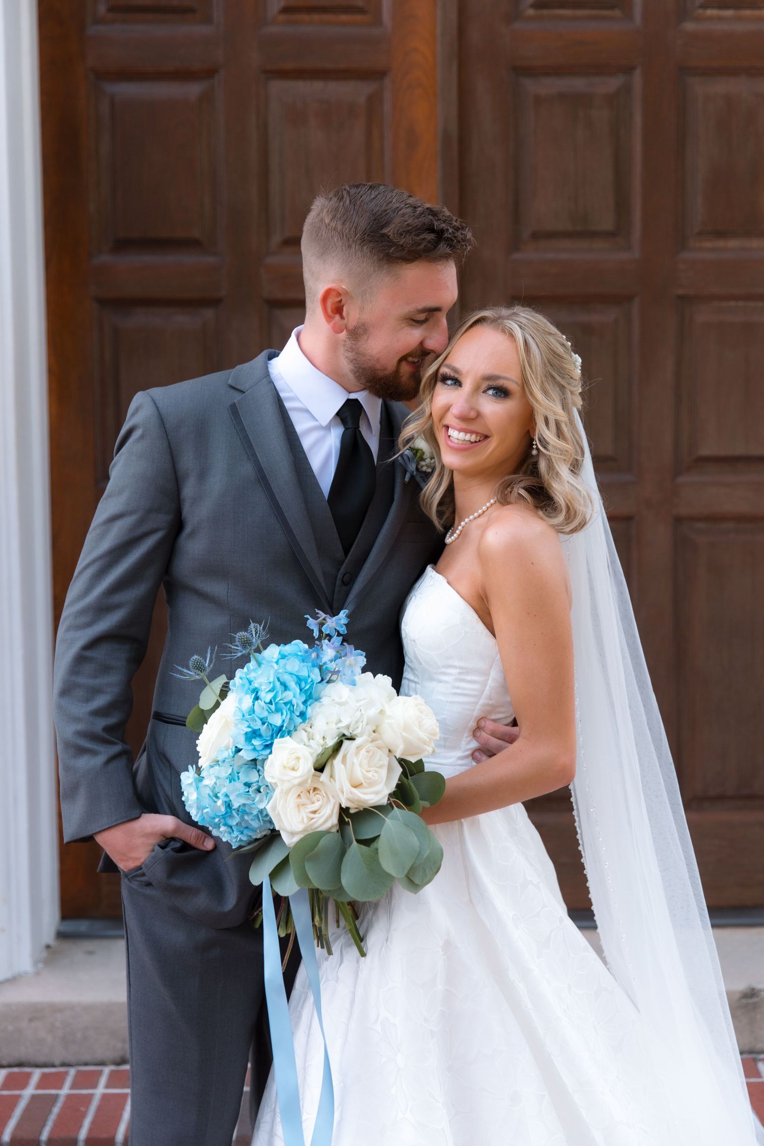 Newlyweds pose together with blue and white floral bouquet in front of wooden church doors.