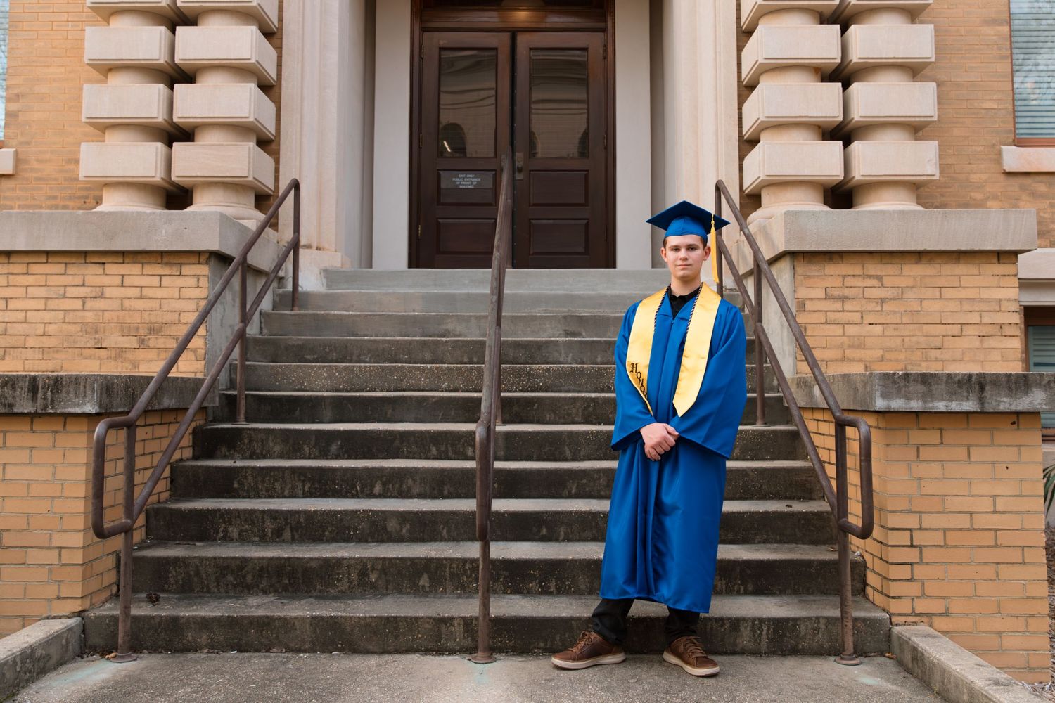 Graduate in blue and gold regalia stands on brick building steps.