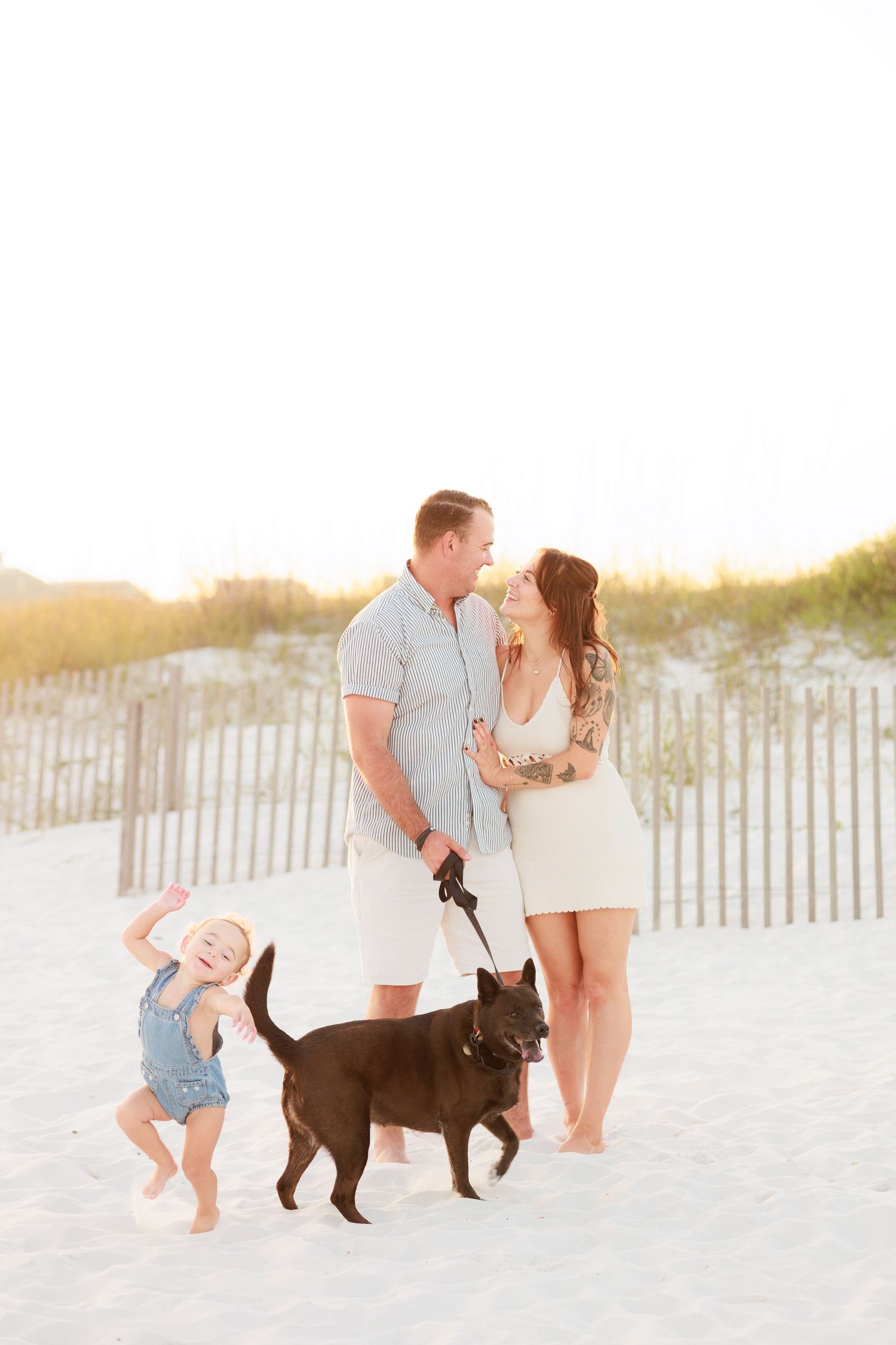 Family beach portrait with couple gazing at each other while toddler dances and dog walks nearby.