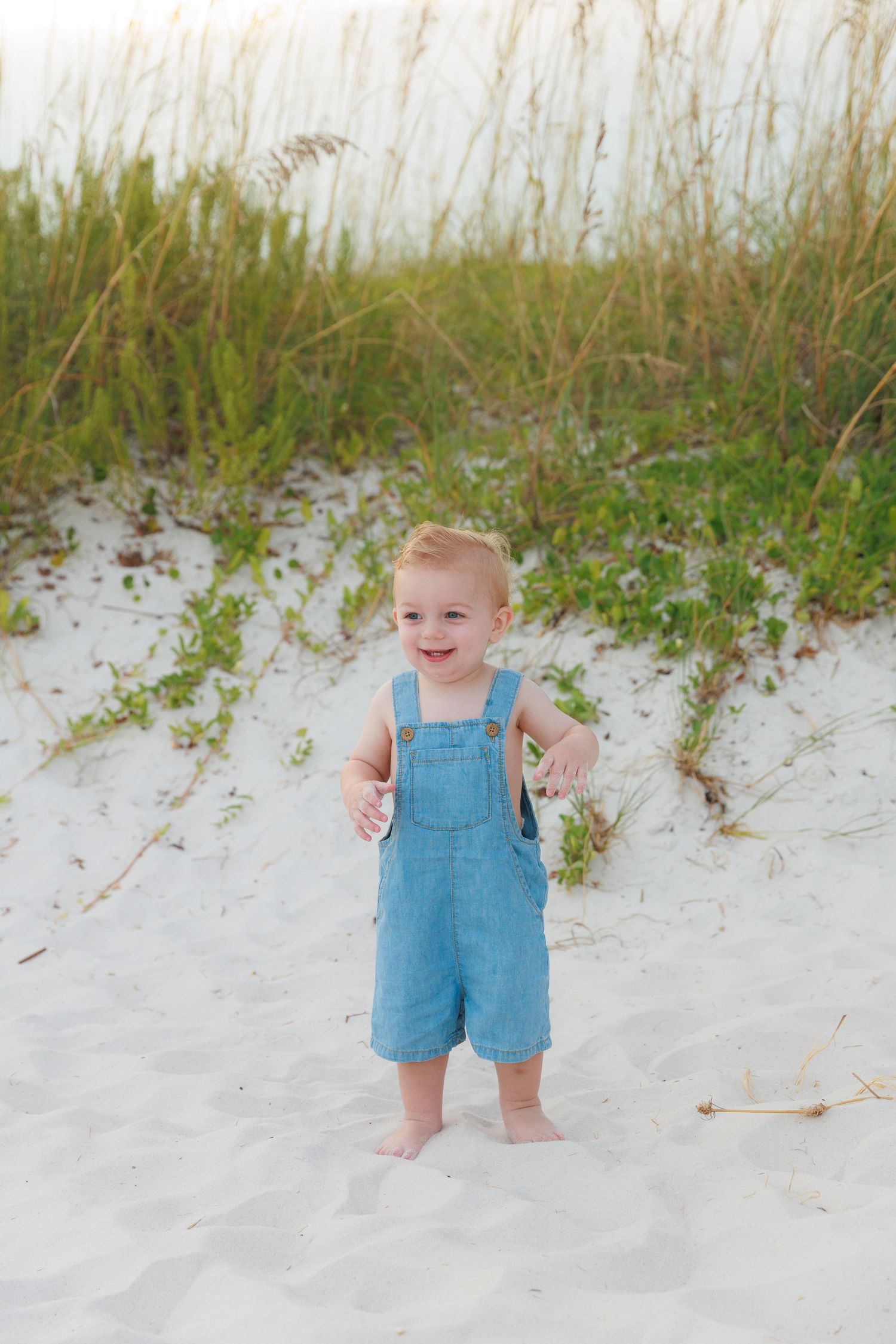 Smiling toddler boy in blue denim overalls standing barefoot on white sand beach near sea oats.