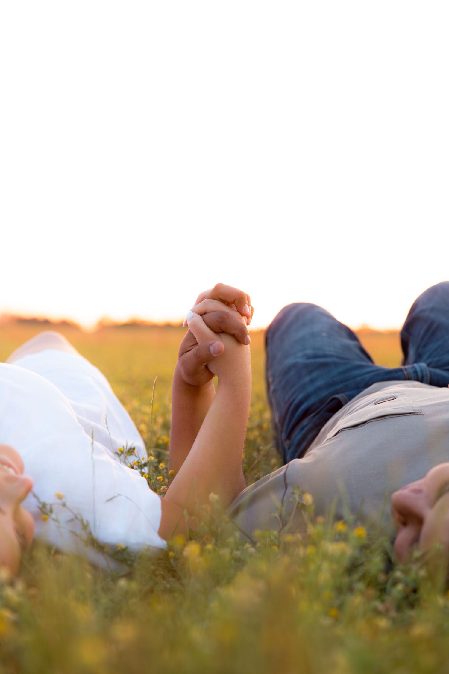 Couple holding hands while lying in a golden wildflower field at sunset, romantic outdoor moment.