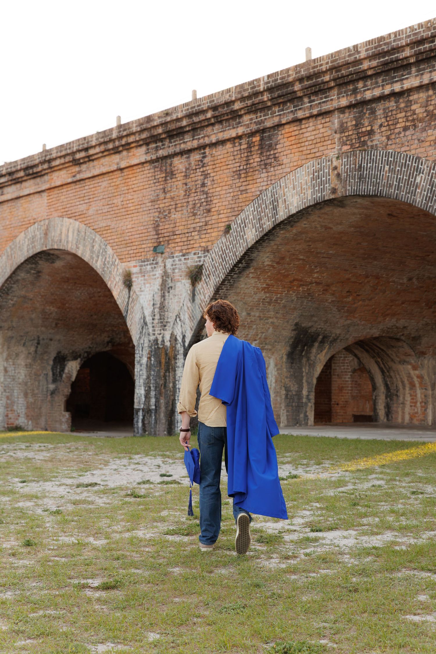 Graduate in blue gown walking away toward brick arched structure.