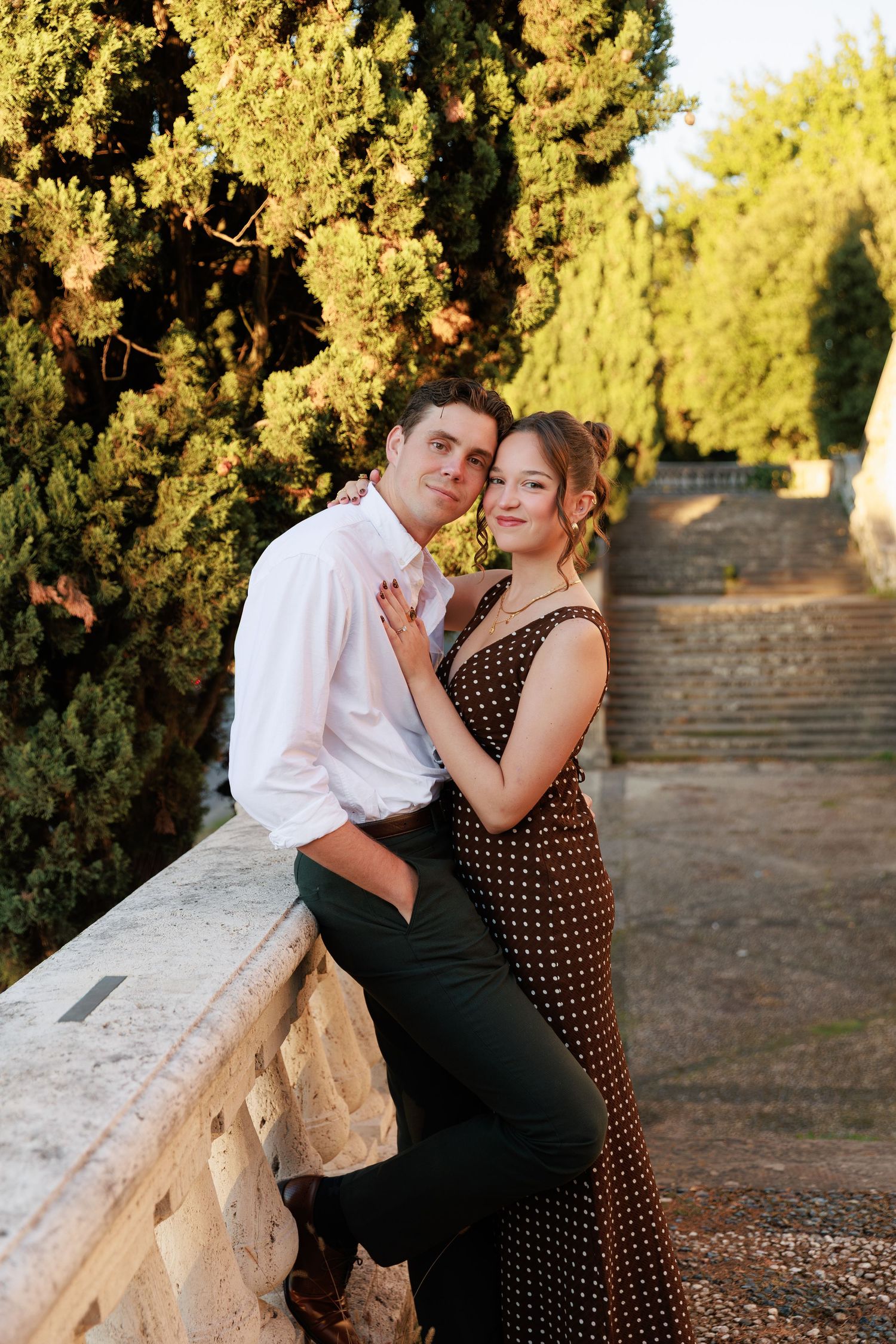 Couple posing romantically against a stone wall surrounded by lush greenery, woman in polka dot dress, man in white shirt.