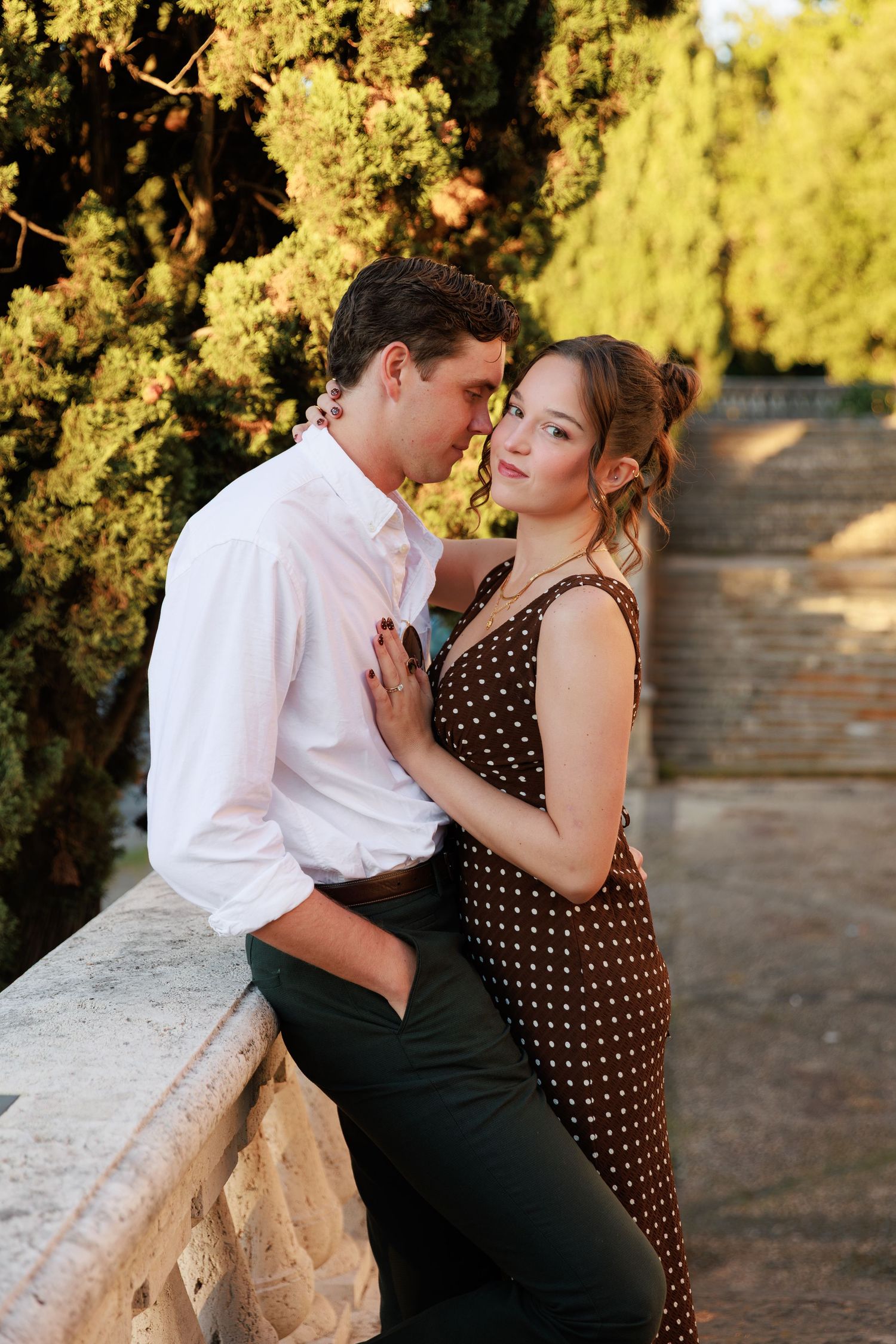 Romantic couple embracing near stone balustrade in sunlit garden, woman in brown polka dot dress gazing at camera.
