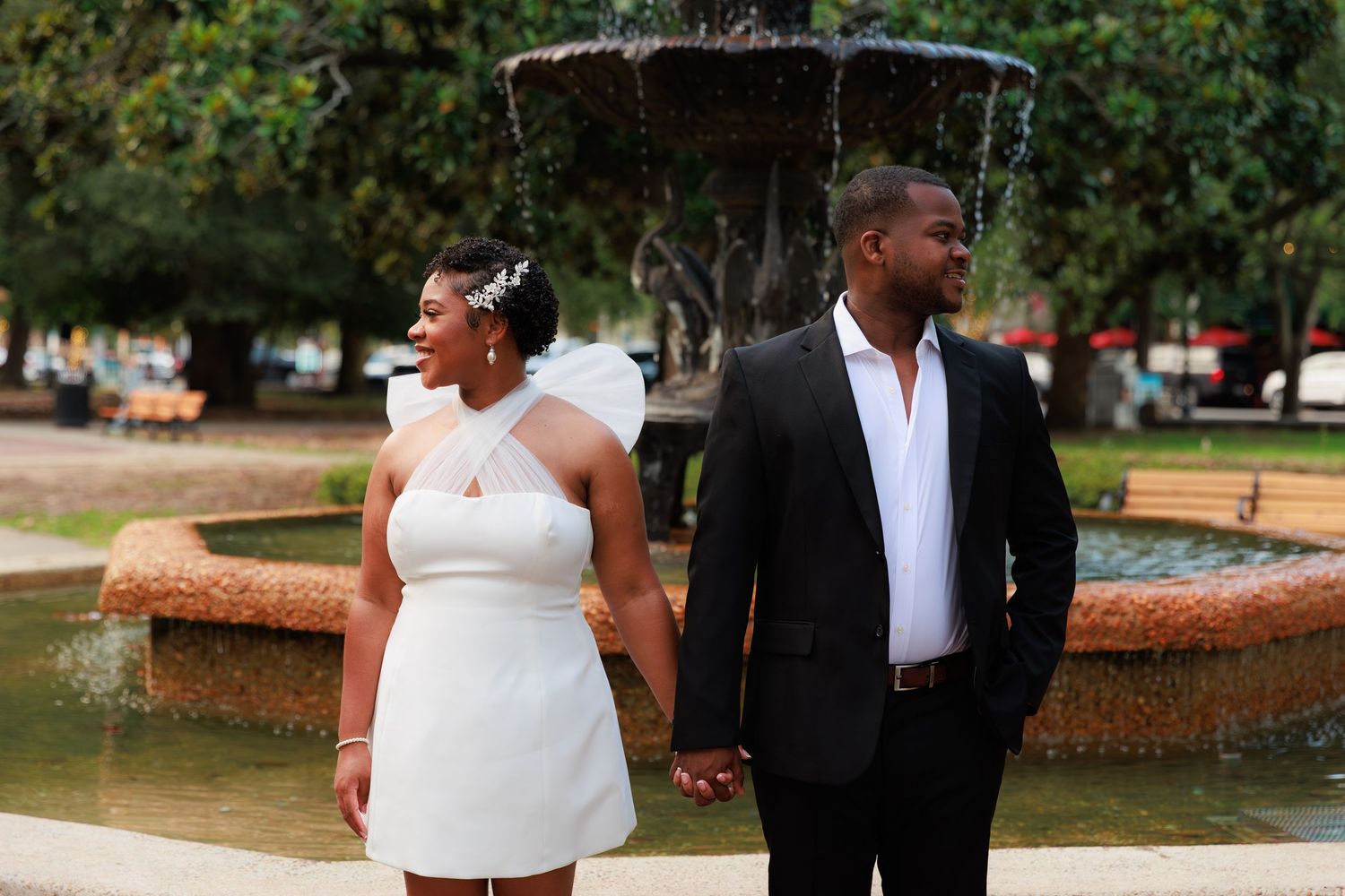 Newlywed couple in wedding attire holding hands by a stone fountain outdoors, bride in white dress, groom in black suit.