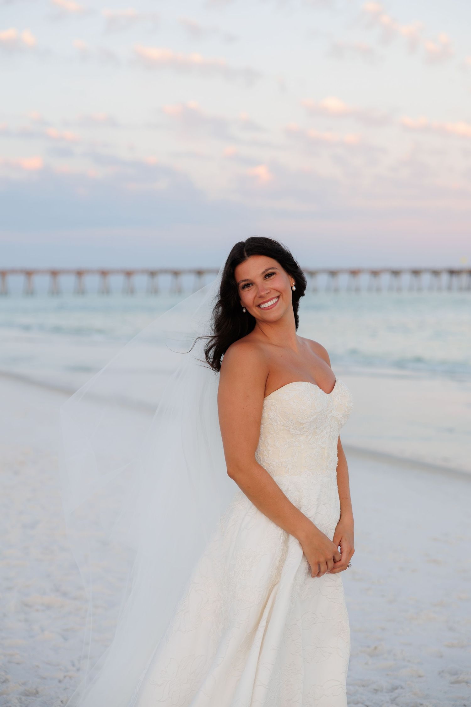 Smiling bride in strapless lace wedding gown posing on white sand beach at sunset with pier in background.
