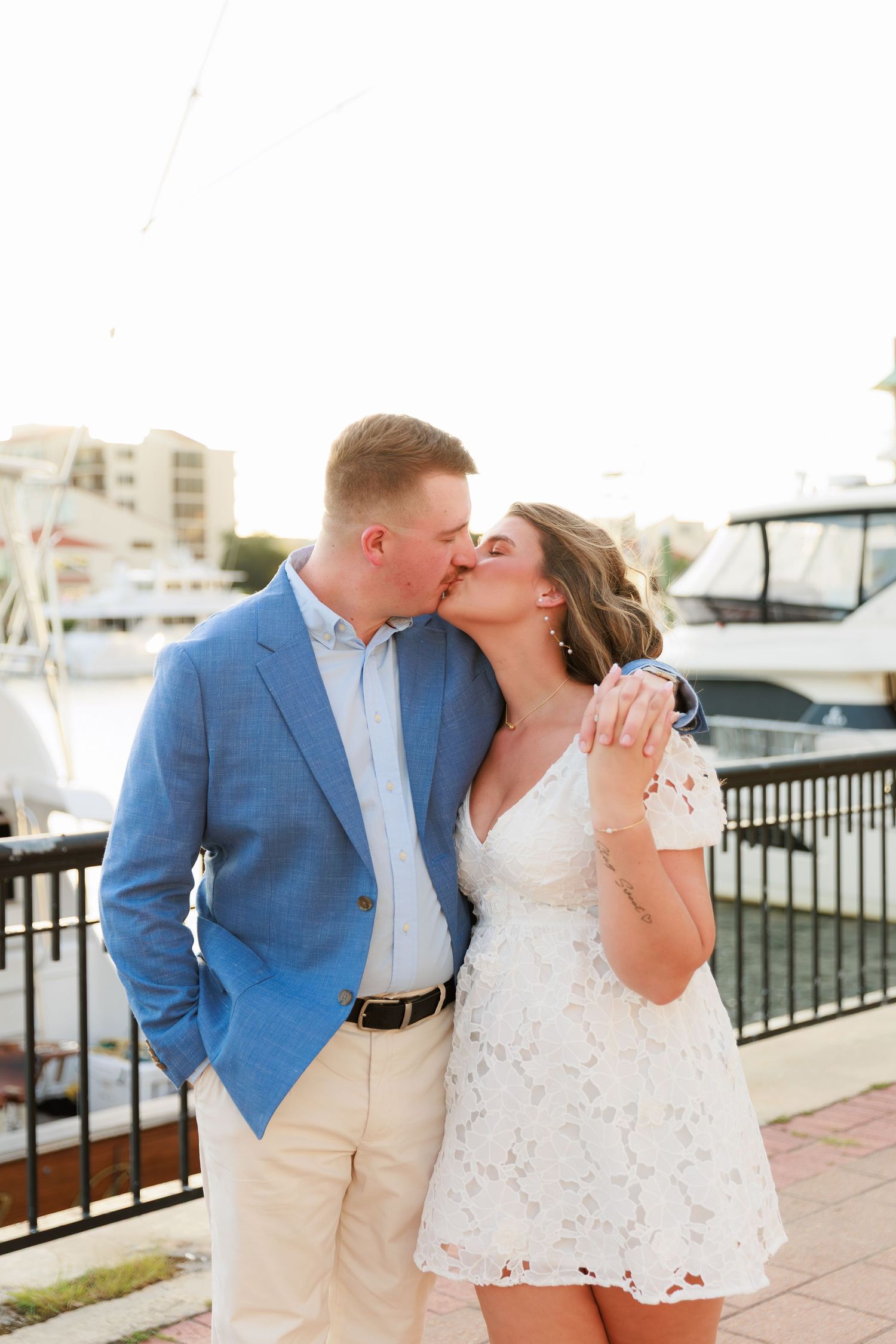 Couple sharing a kiss near a marina at sunset, man in blue blazer and woman in white lace dress.