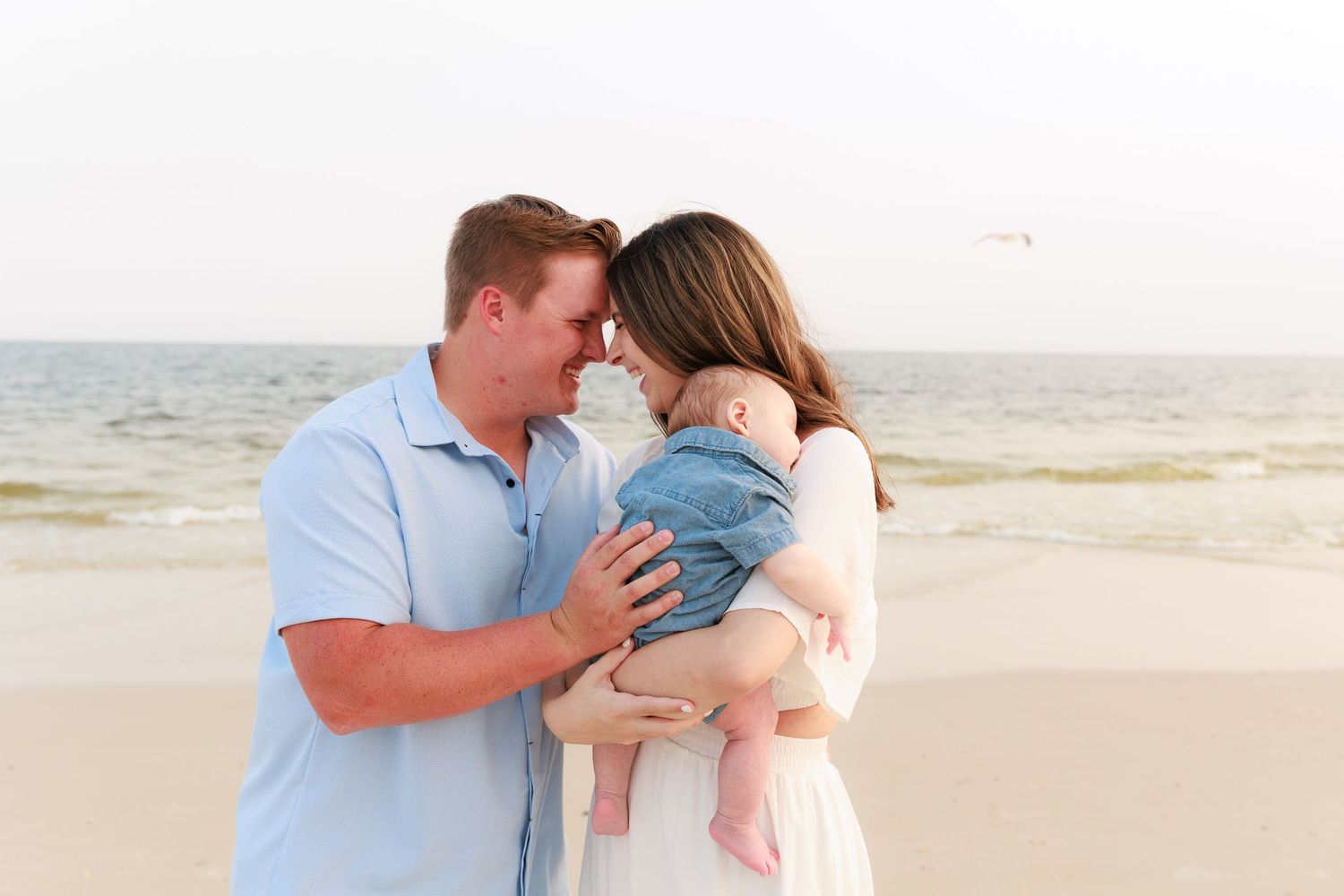 Young couple sharing tender moment on beach while holding their infant baby dressed in denim.