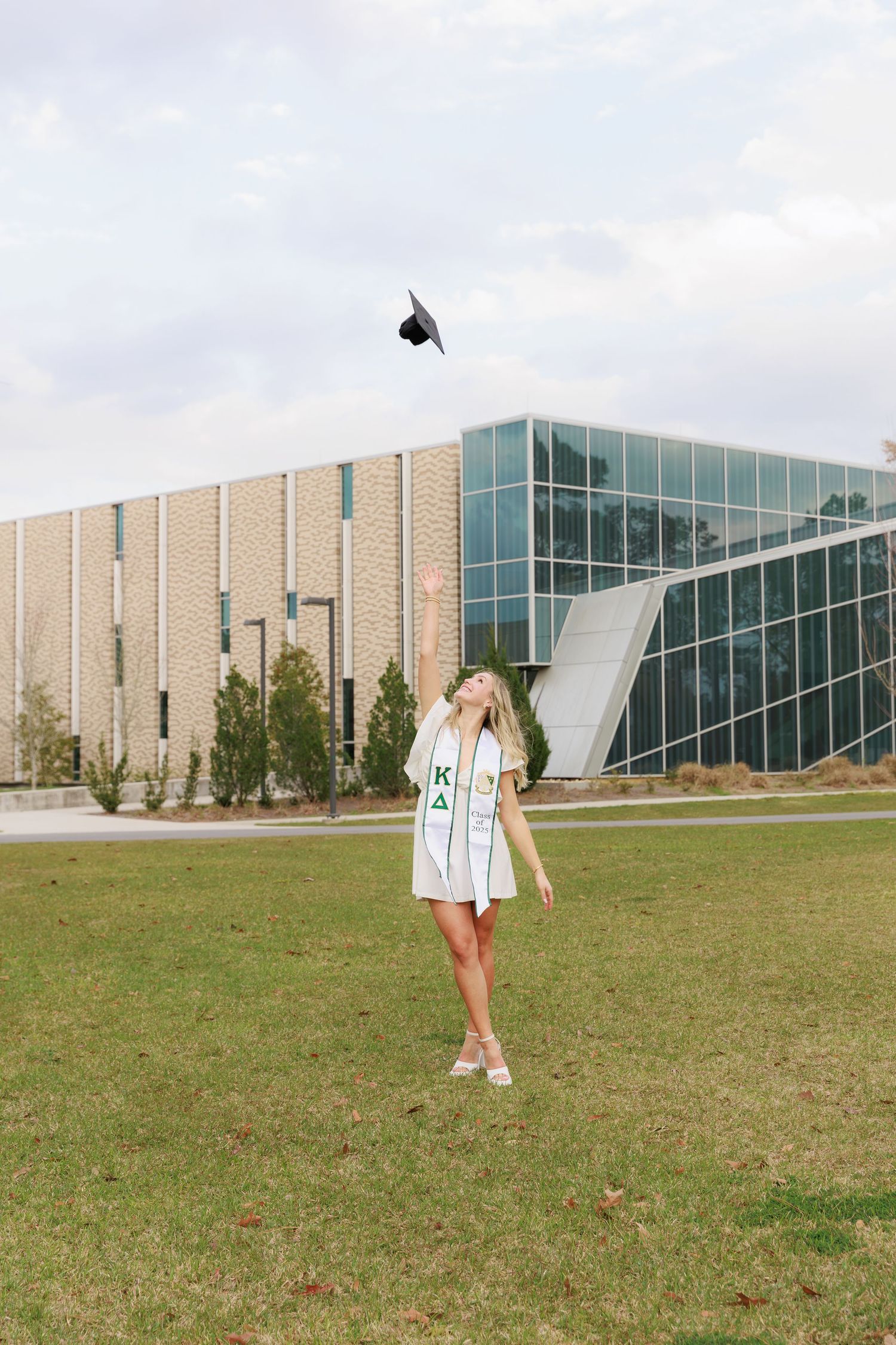 College graduate tosses cap in air wearing Kappa Delta sash, celebrating outdoors near modern university building.