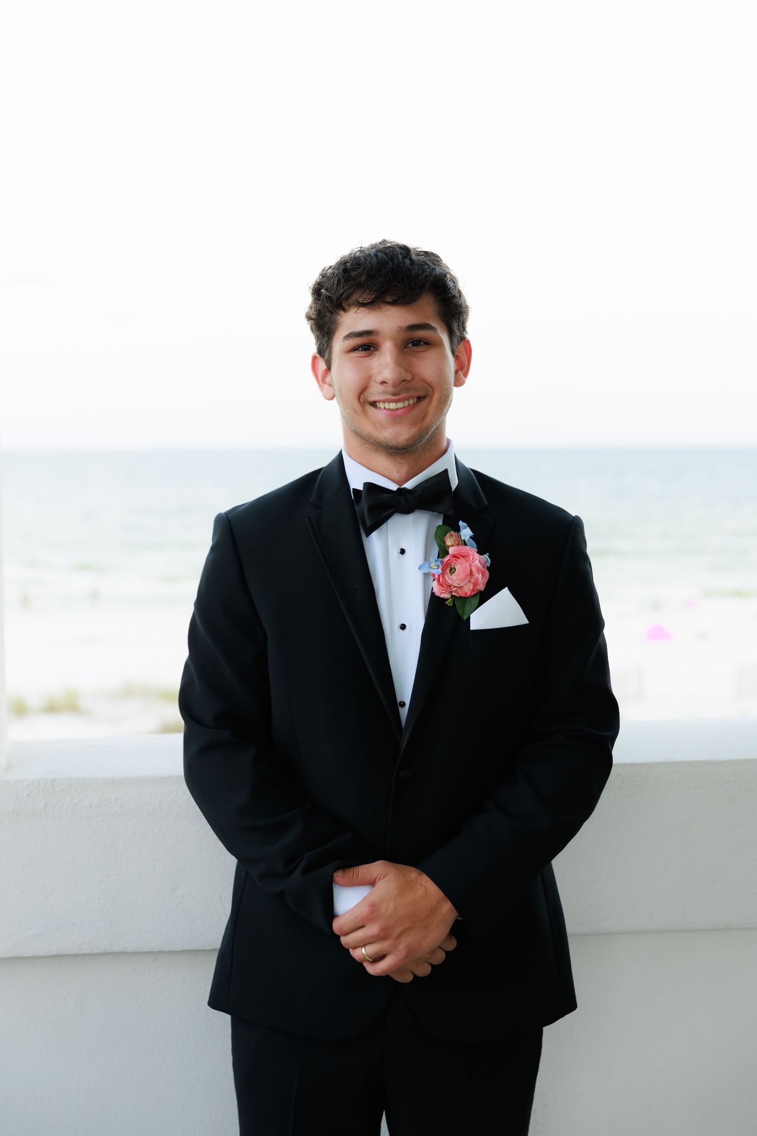 A groom in a black tuxedo with coral boutonniere smiles at a beach wedding venue.