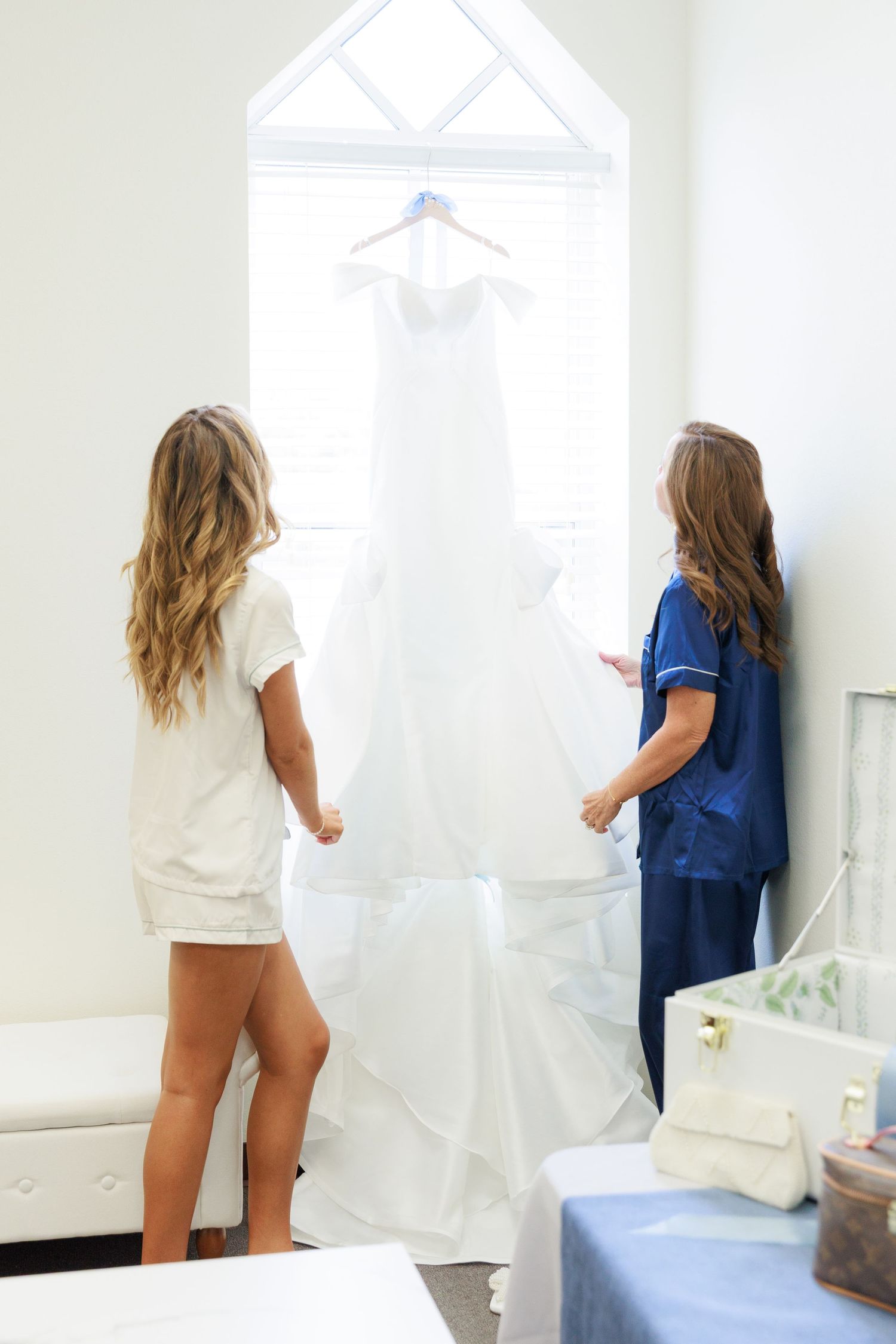 Two people in a room admire a white wedding dress hanging in front of a bright window.