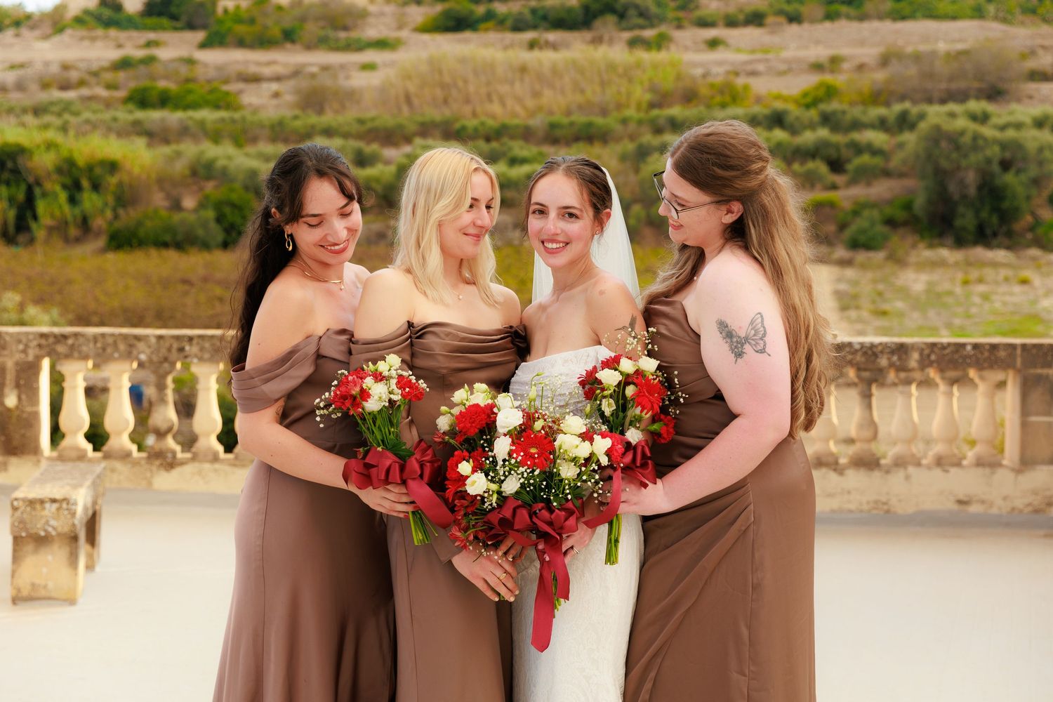 Bridesmaids in chocolate brown dresses stand together holding vibrant red and white bouquets at an outdoor venue.