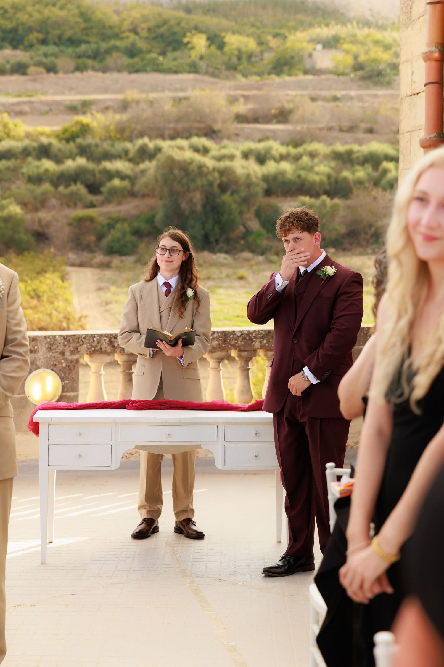 A sequence of wedding guests sitting on a stone wall at sunset during an outdoor reception.