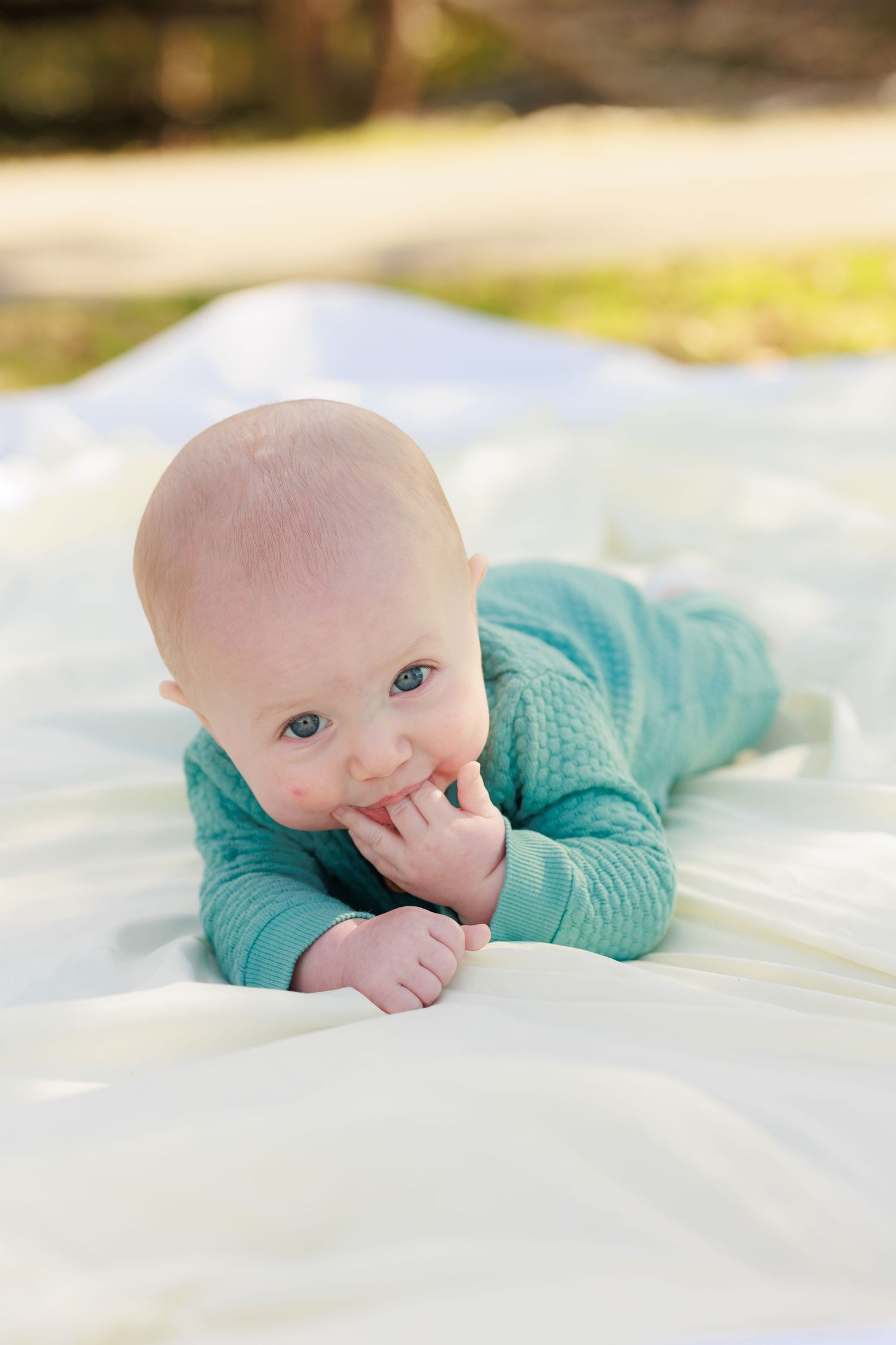 Baby in turquoise sweater lying on white blanket with hand near mouth in outdoor setting.