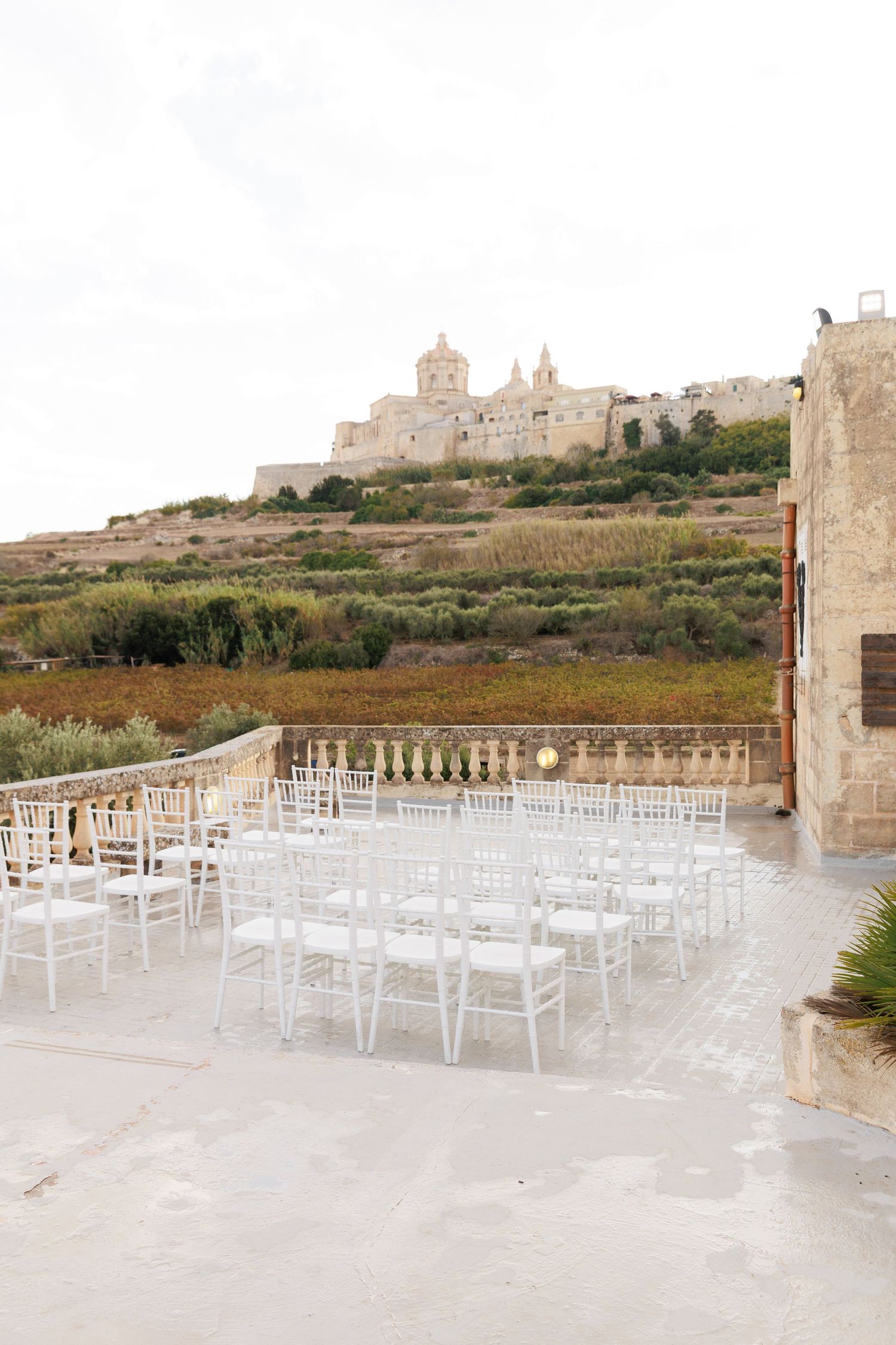 White chairs arranged for an outdoor wedding ceremony with a historic hilltop cathedral visible in the background.
